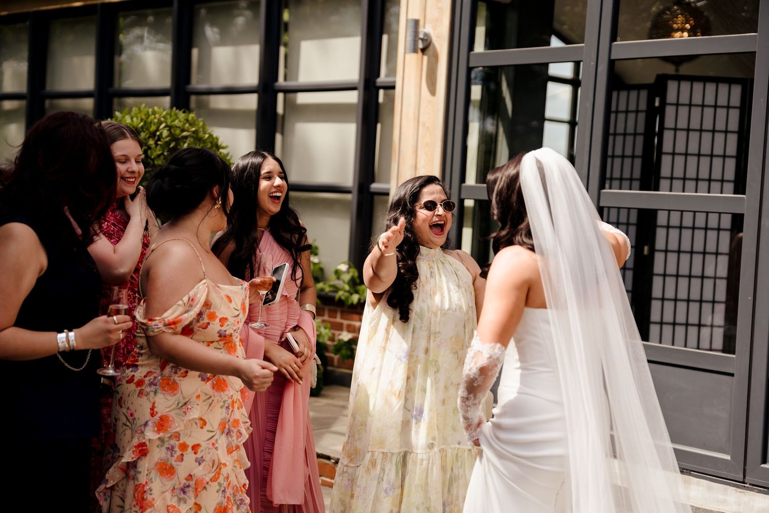 Bride in white gown and veil laughing with bridesmaids in colorful dresses at outdoor wedding venue.