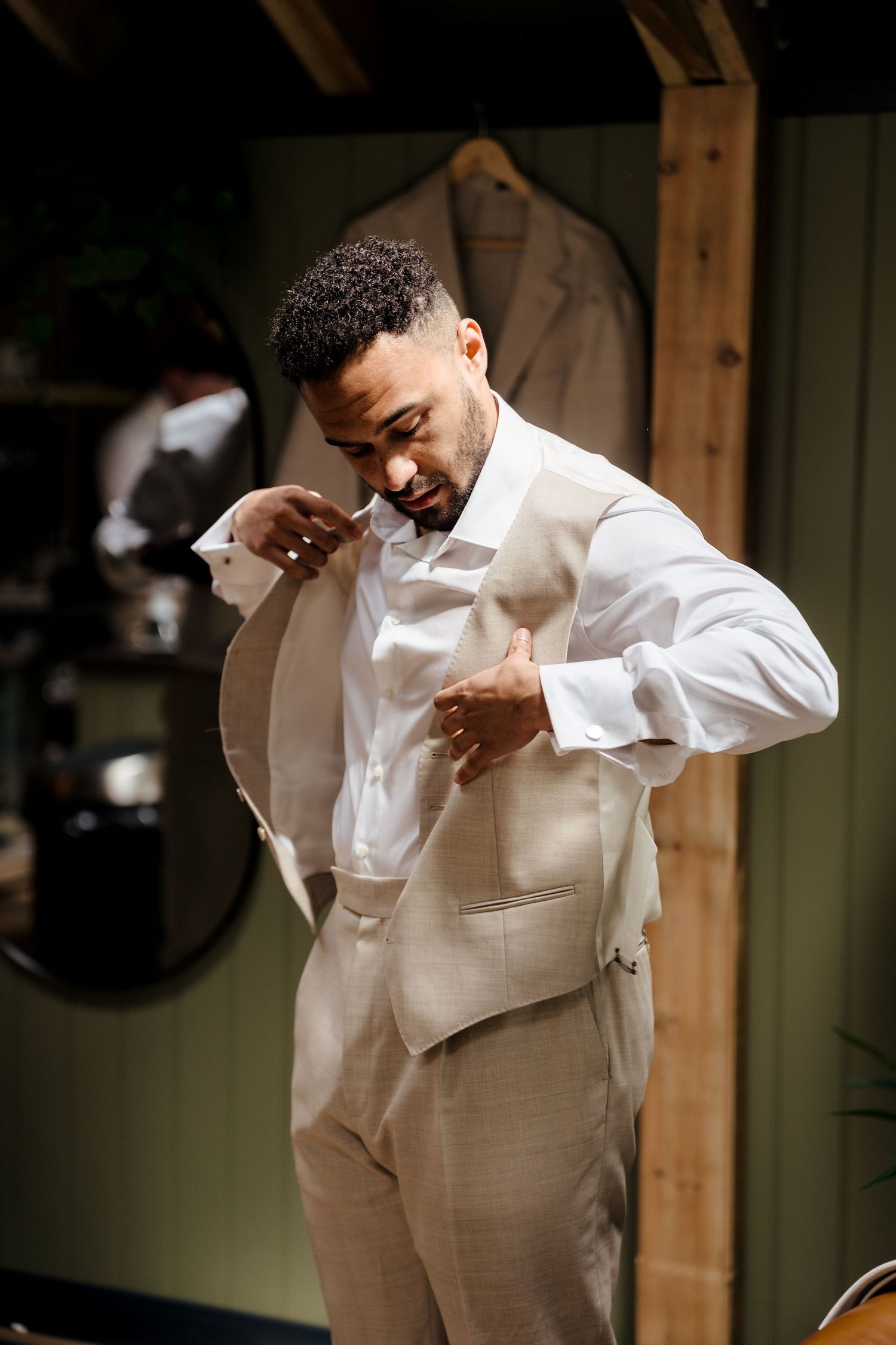 Groom in white shirt and beige vest getting dressed for his wedding day in a rustic dressing room.