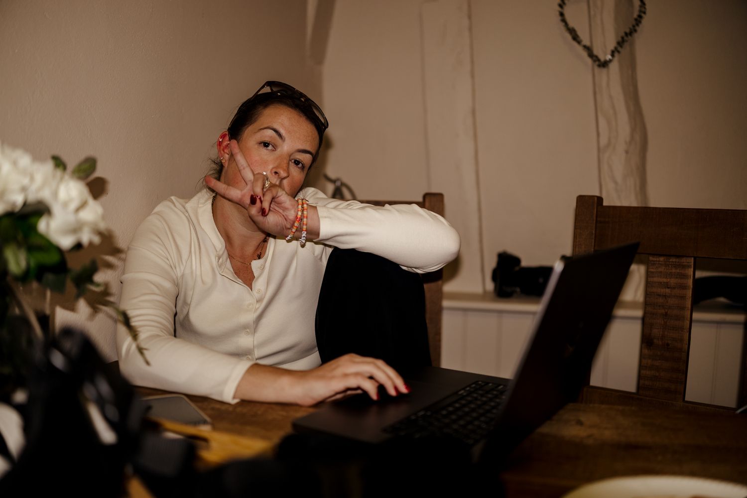 Woman sitting at wooden table working on laptop, gesturing with hand, in cozy dimly lit home office.