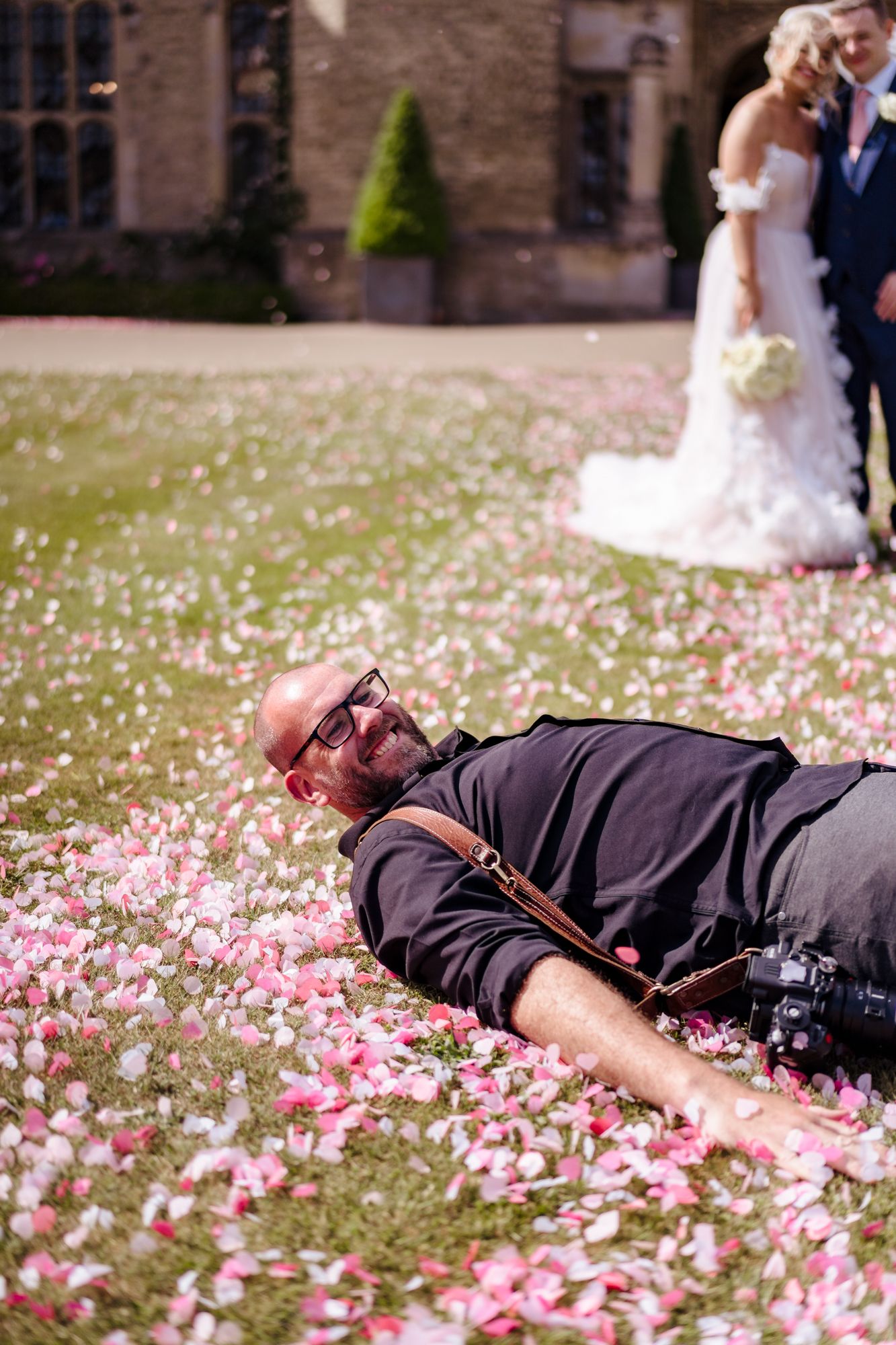 A wedding photographer lies on flower petal covered grass capturing creative low angle shots of a bride and groom.