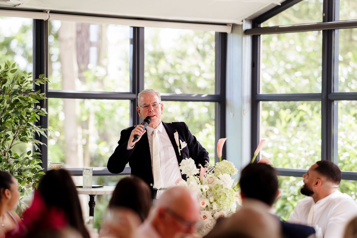 Smiling man in black suit delivers speech at microphone during elegant indoor wedding reception.