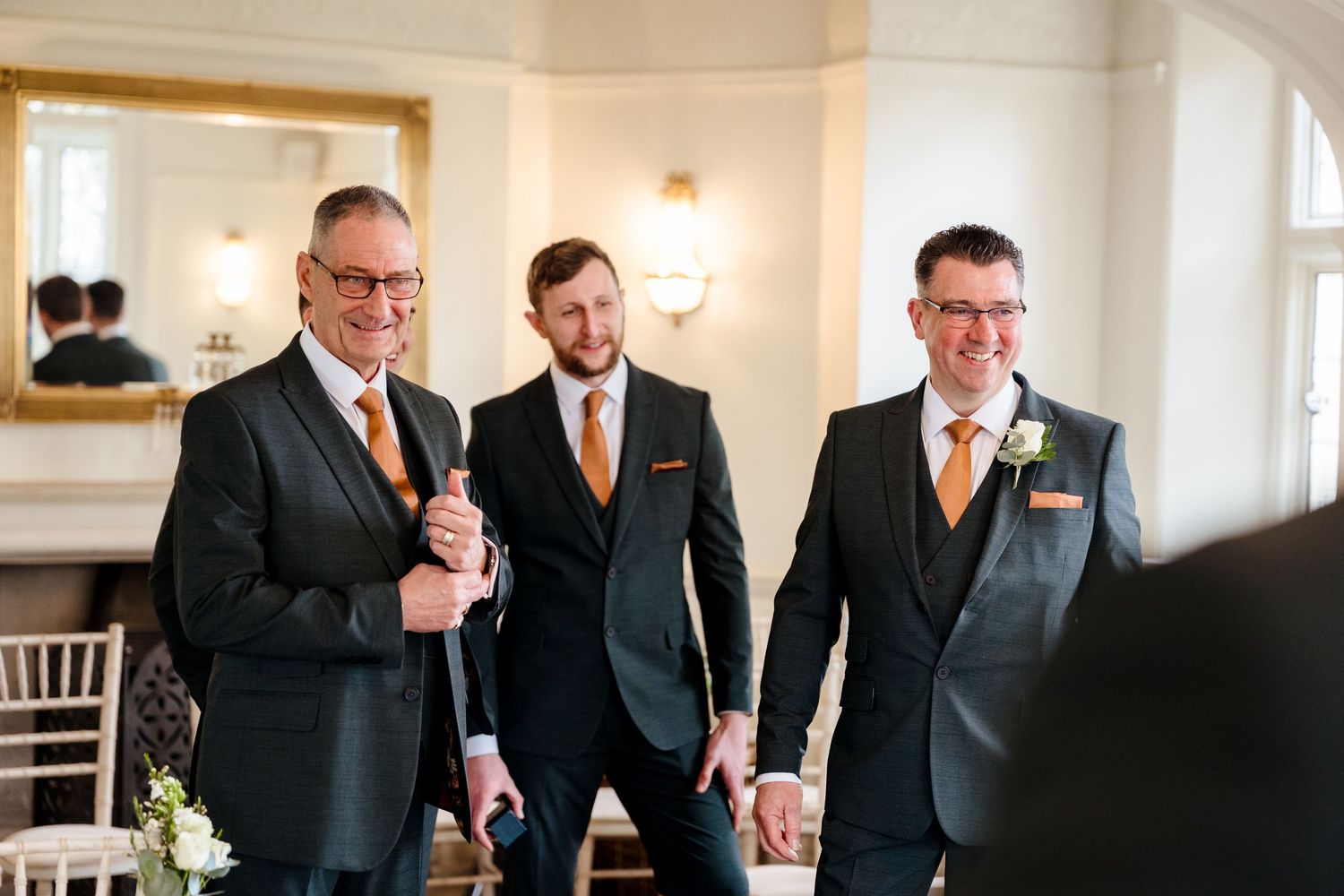 Three groomsmen in dark suits with orange ties smiling together in an elegant wedding venue.