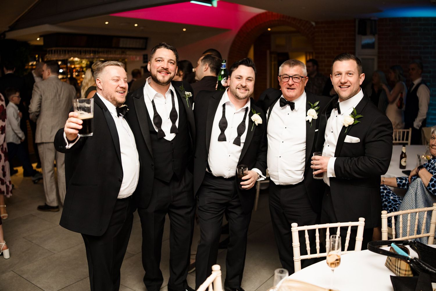 Five groomsmen in black tuxedos raising glasses and smiling at elegant wedding reception venue.
