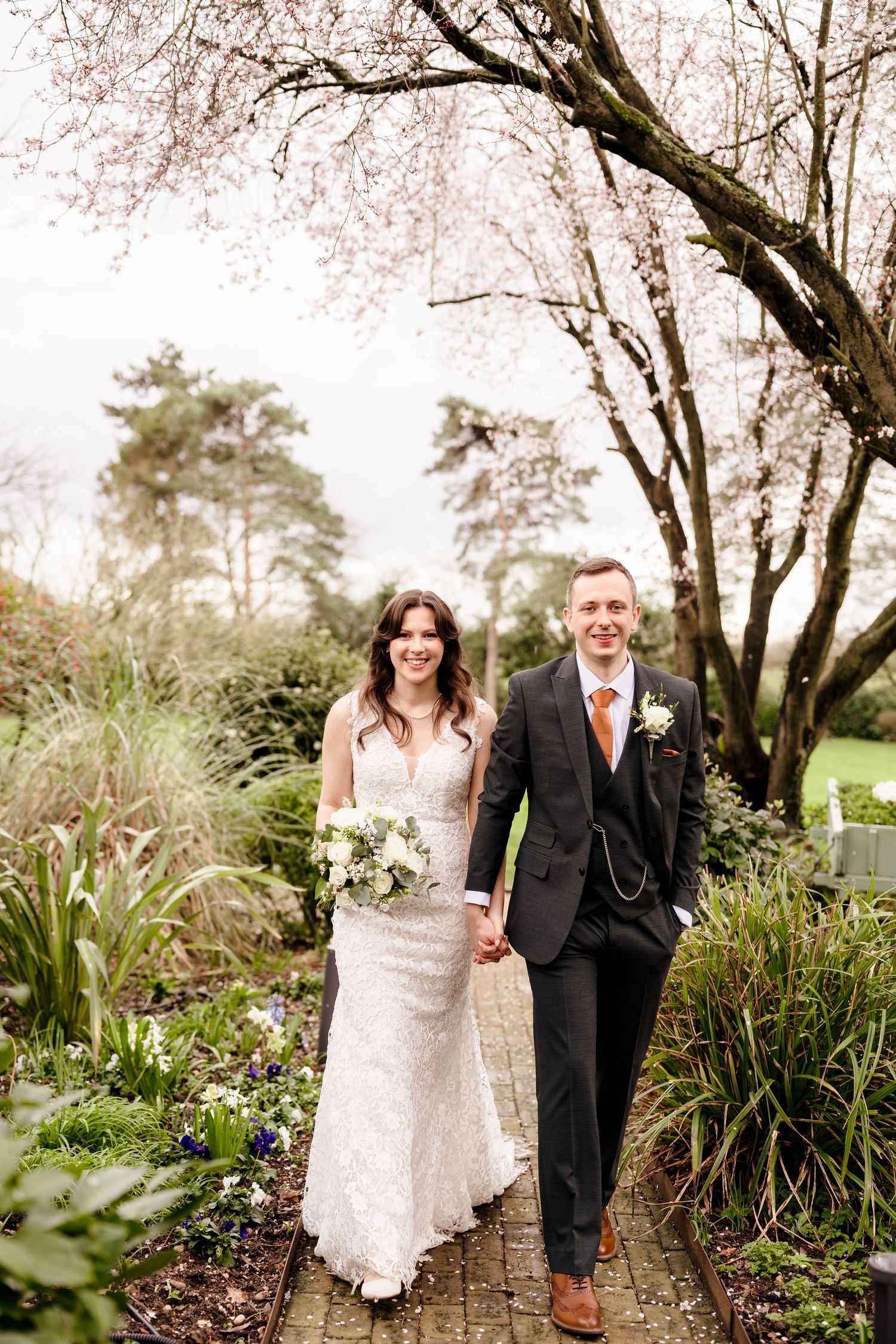 Bride and groom walk hand-in-hand through a garden path under cherry blossom trees on their wedding day.
