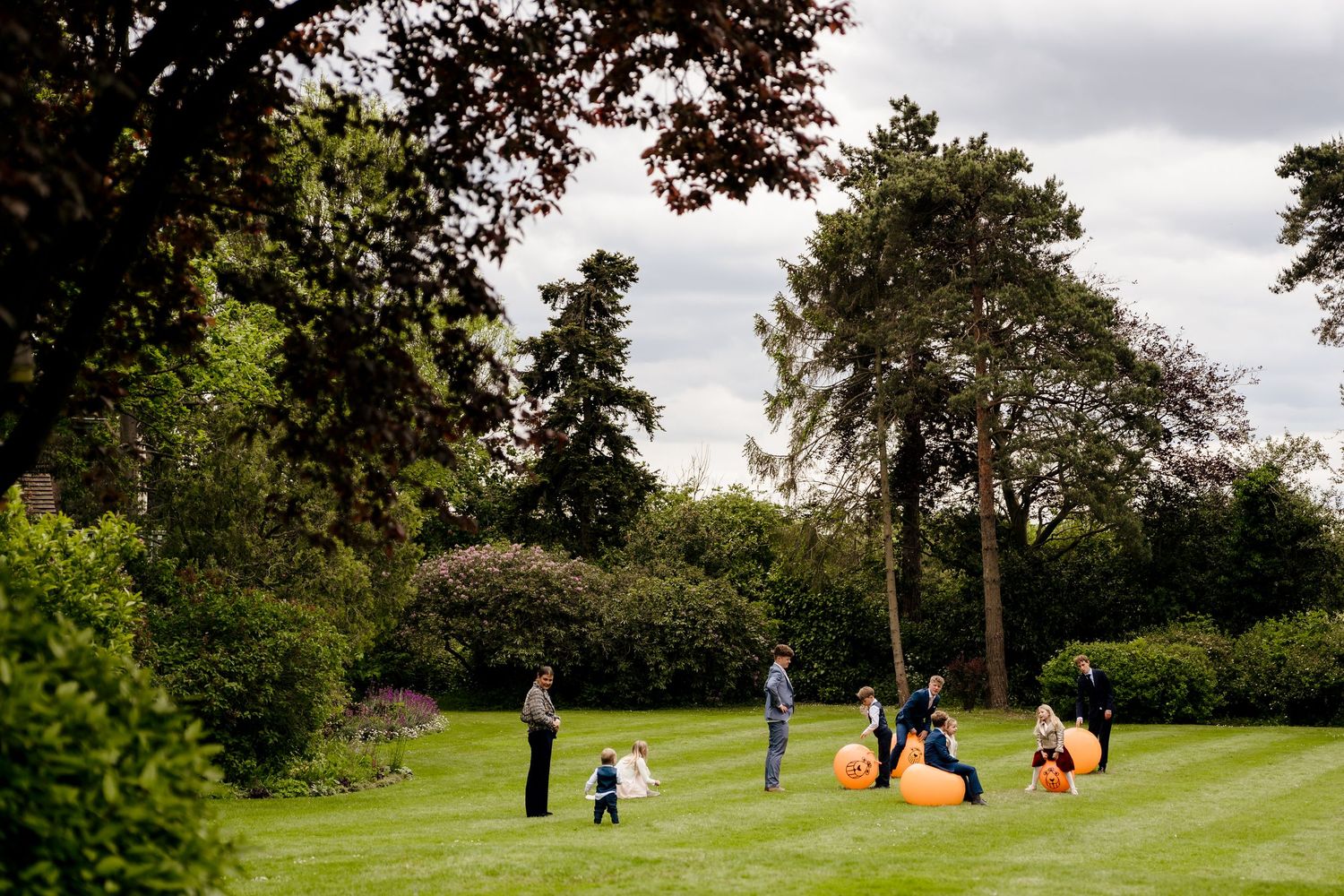 Children playing on orange bean bags in a lush green garden surrounded by tall trees on a cloudy day.