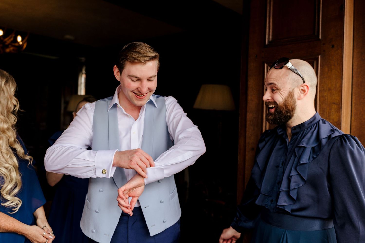 Groom adjusting cufflinks with groomsmen smiling in elegant wedding preparation setting.