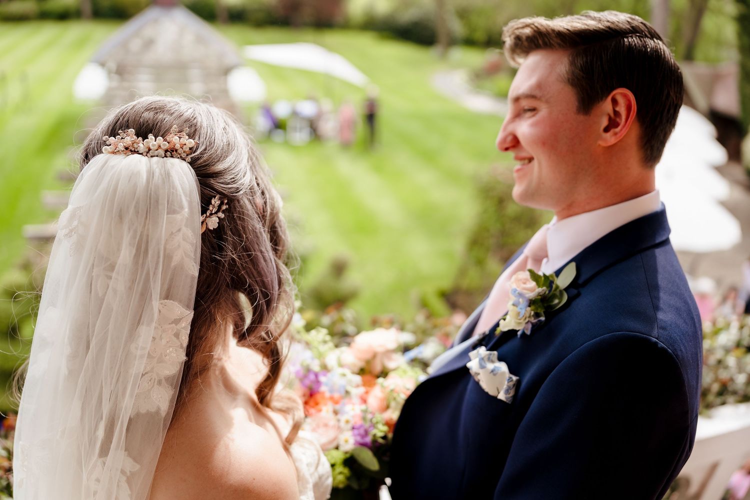 Smiling groom in navy suit gazes at veiled bride during outdoor garden wedding ceremony.