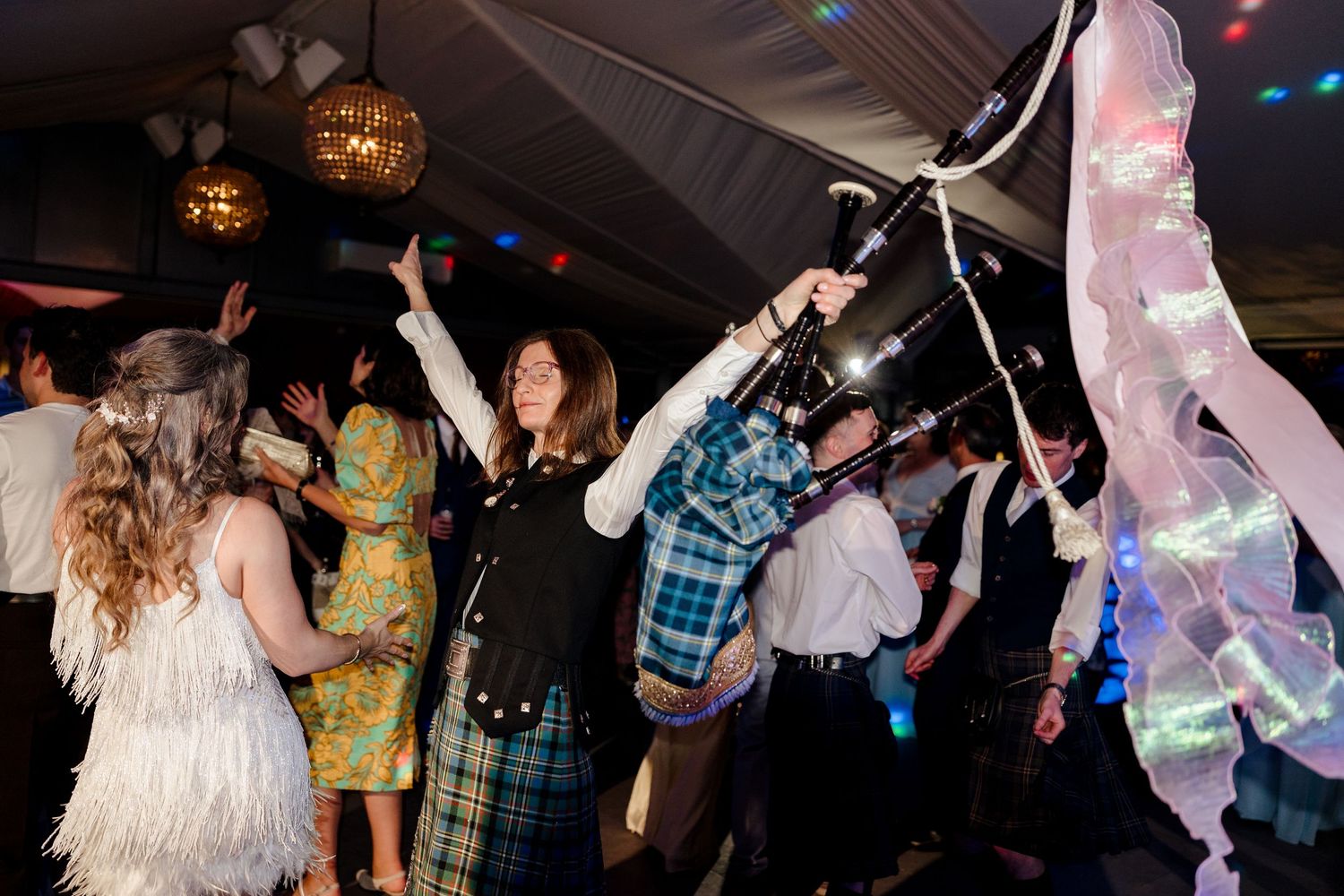 Lively wedding reception dance floor with bagpiper in tartan and guests dancing under festive lights.