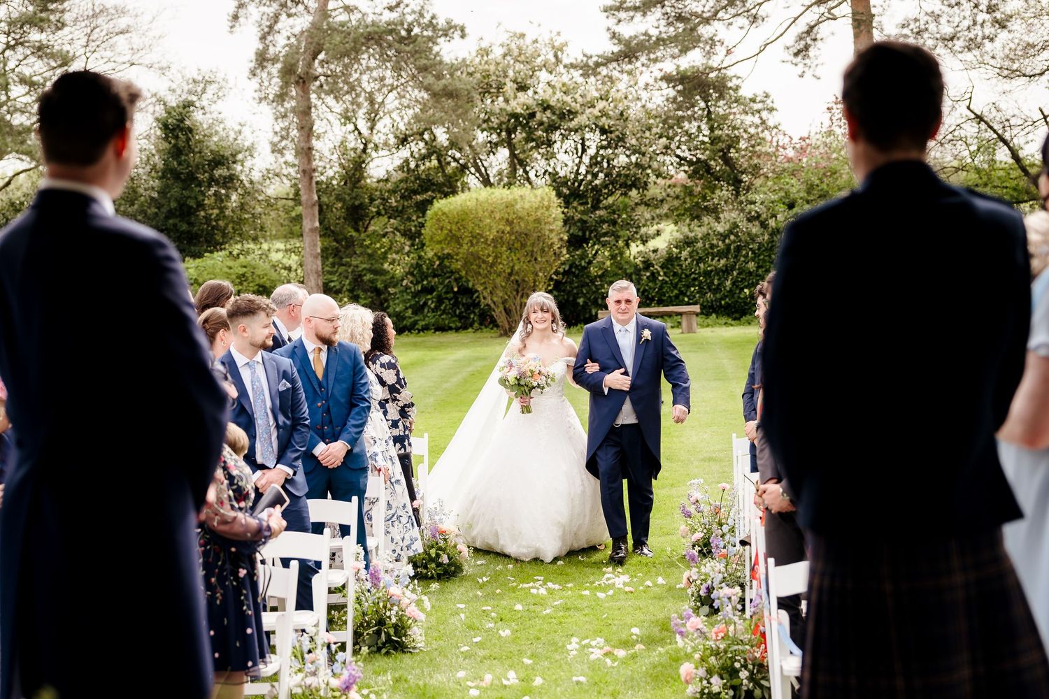 Bride walks down outdoor aisle in white gown with escort at elegant garden wedding ceremony.