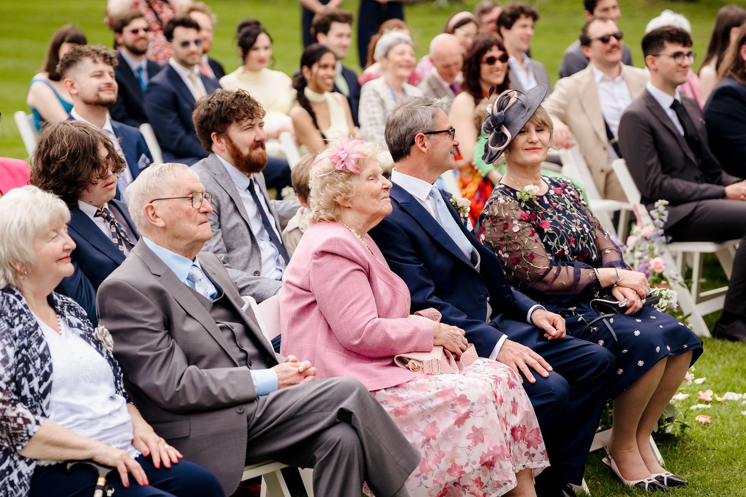 Wedding guests seated outdoors in white chairs smiling during a sunny garden ceremony.