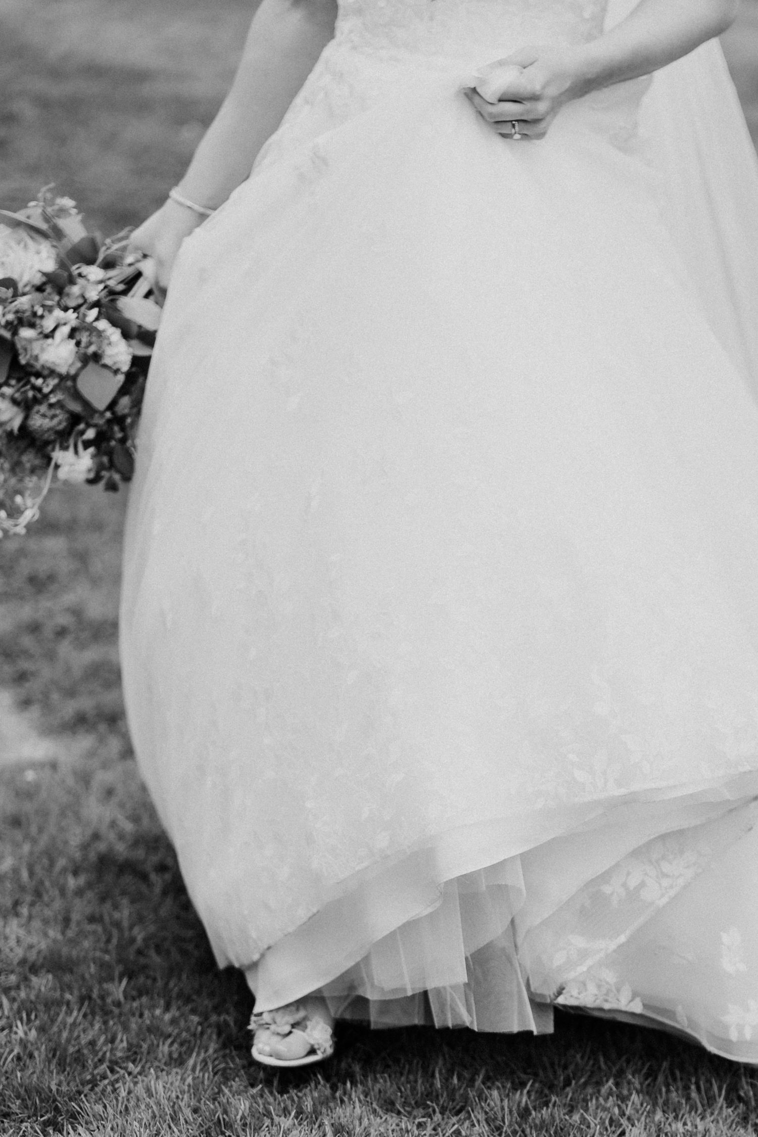 Black and white close-up of bride walking in flowing lace wedding gown holding a floral bouquet.