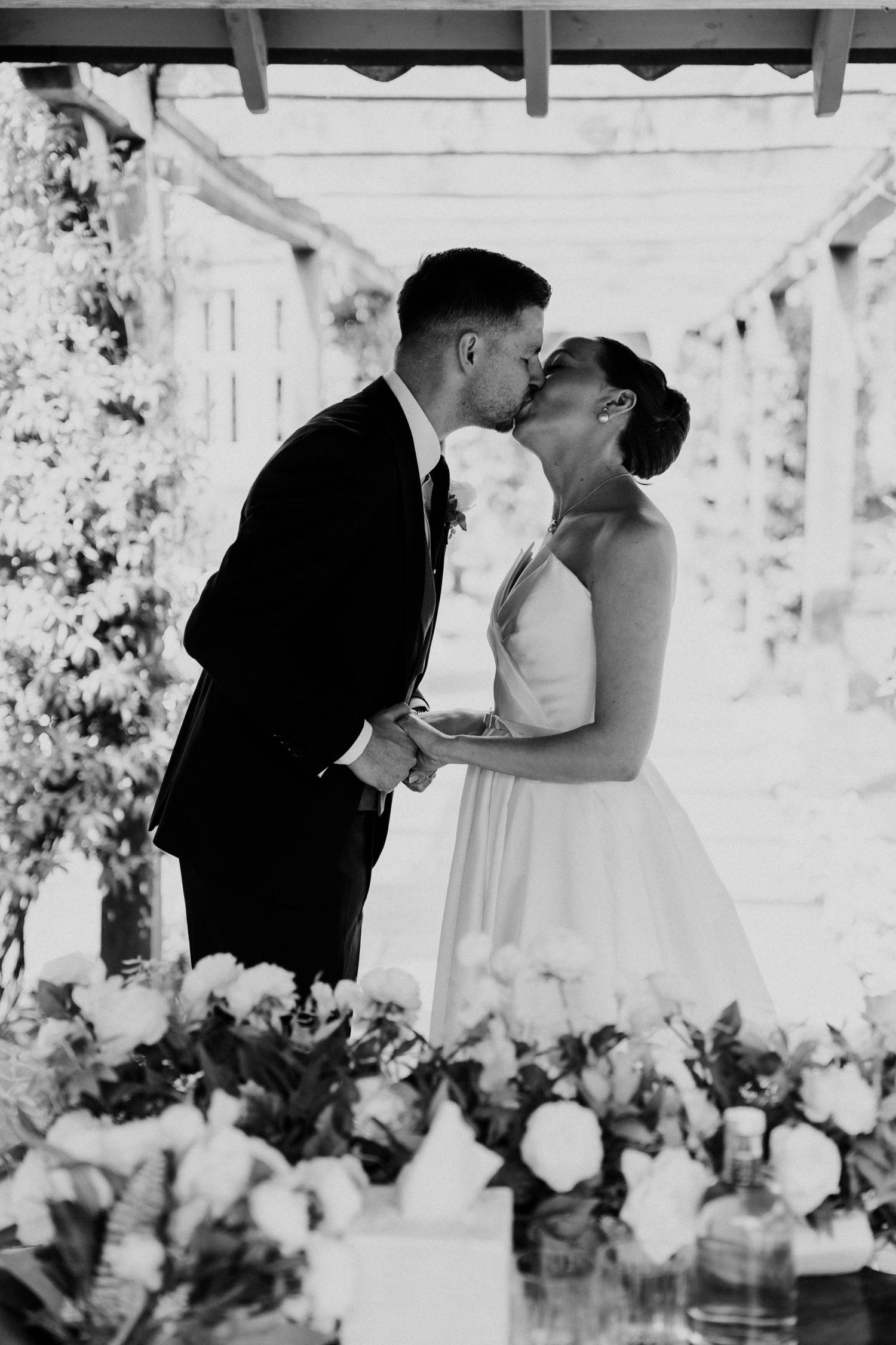 Black and white photo of bride and groom sharing first kiss at floral-decorated wedding altar.