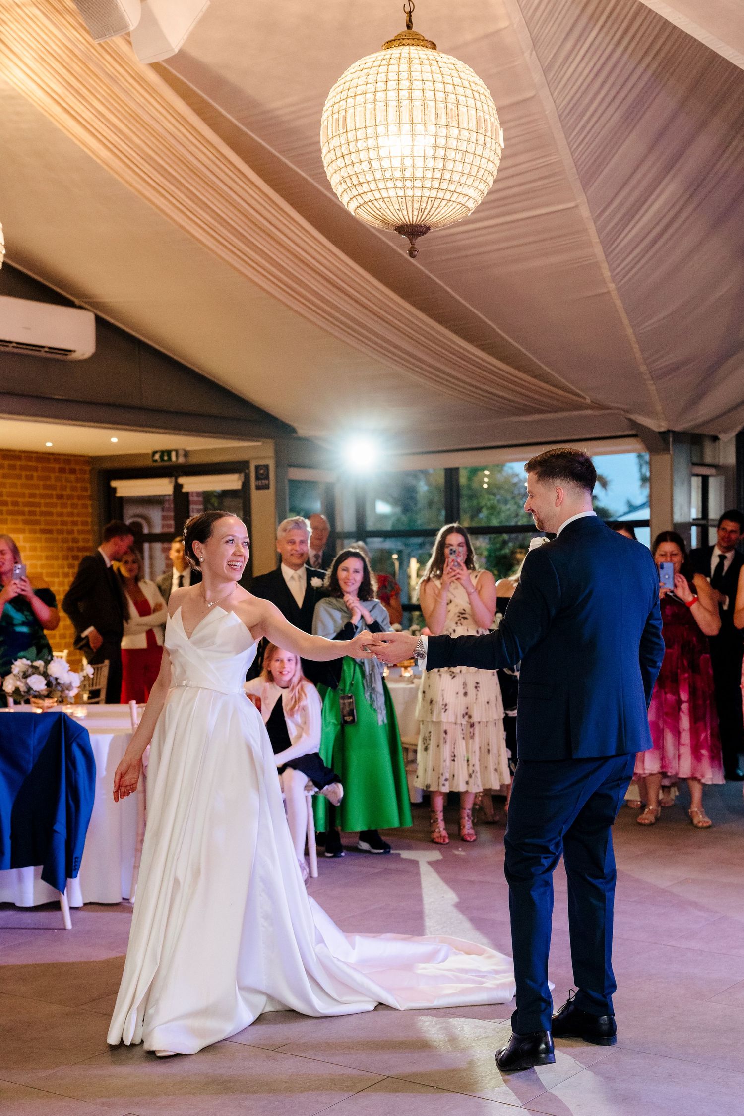 A bride and groom share their first dance under a glowing chandelier surrounded by guests.