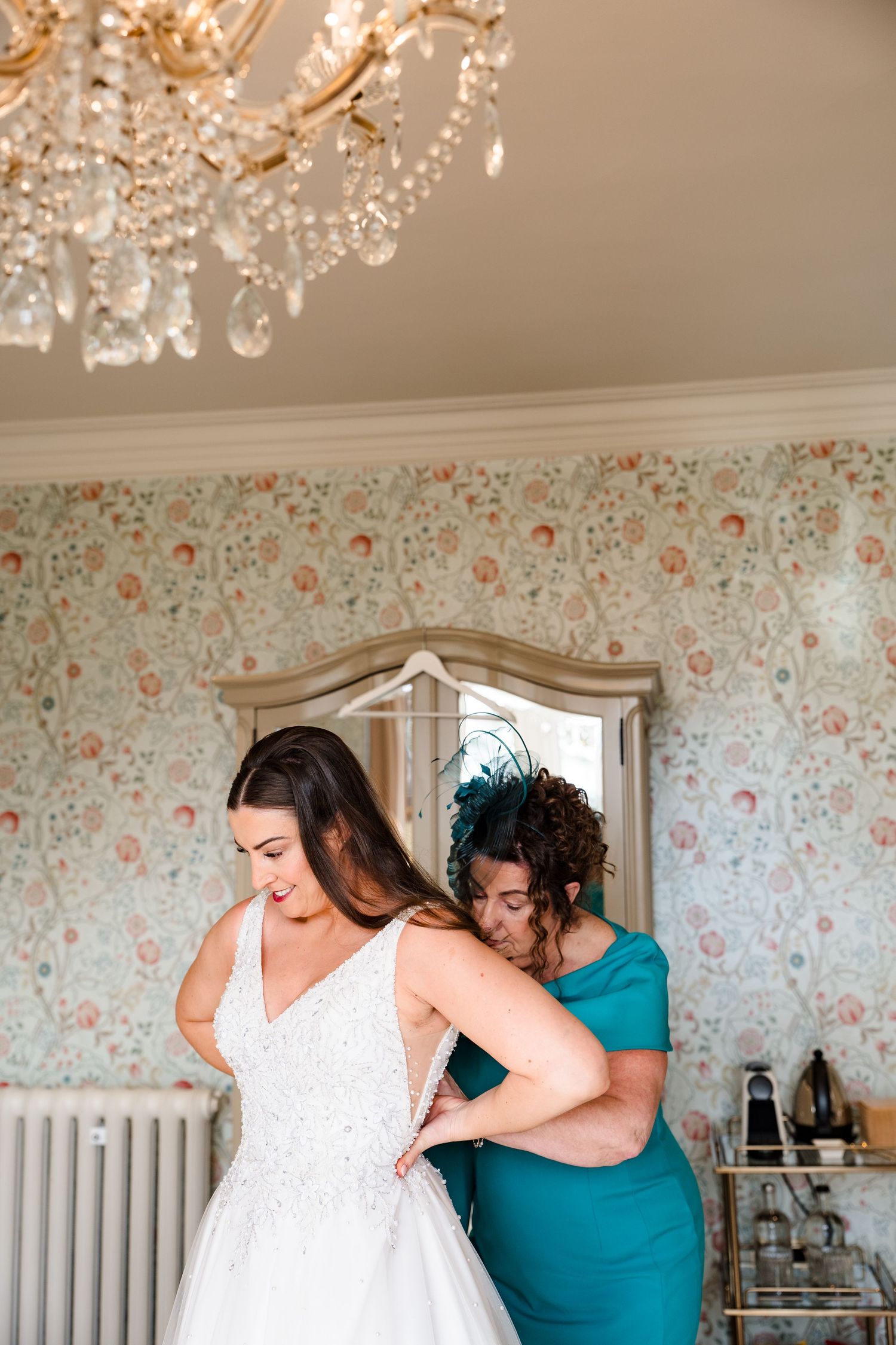Bridesmaid in teal dress helps bride with her white lace wedding gown in a floral wallpapered room.
