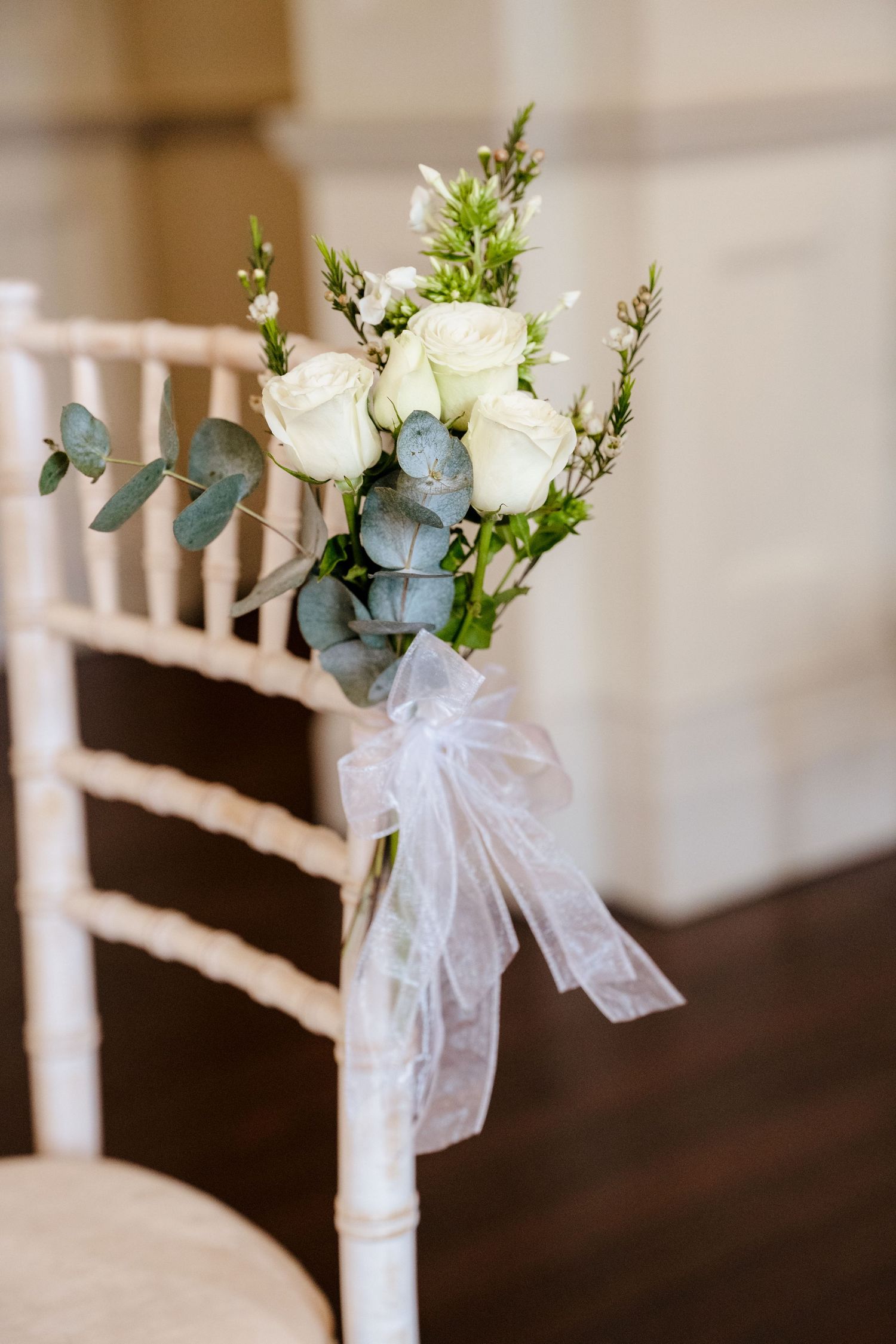 White roses and eucalyptus wedding chair decoration tied with sheer ribbon bow at indoor ceremony.