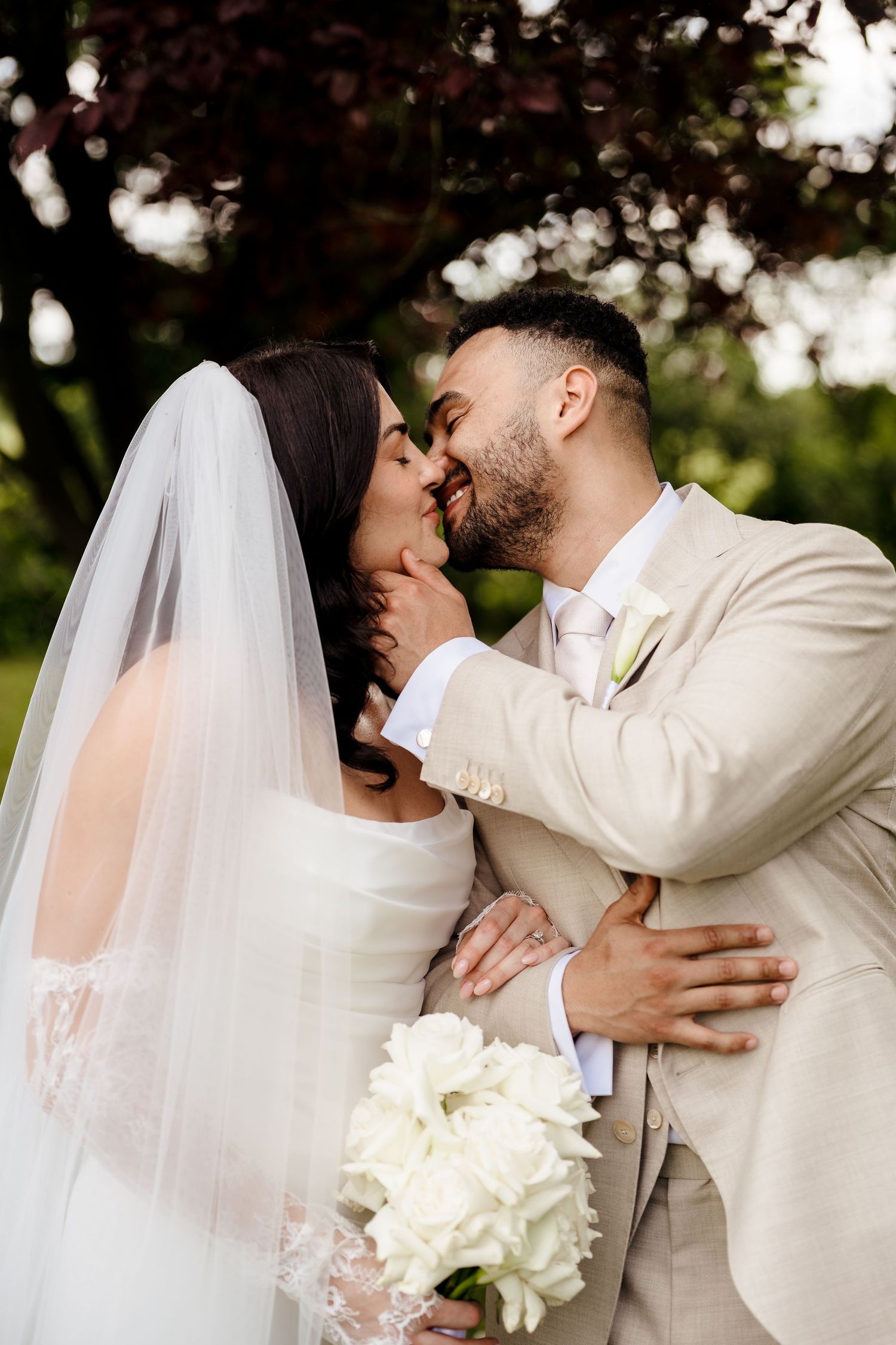 Bride and groom share romantic kiss outdoors, bride holding white calla lily bouquet under leafy tree.