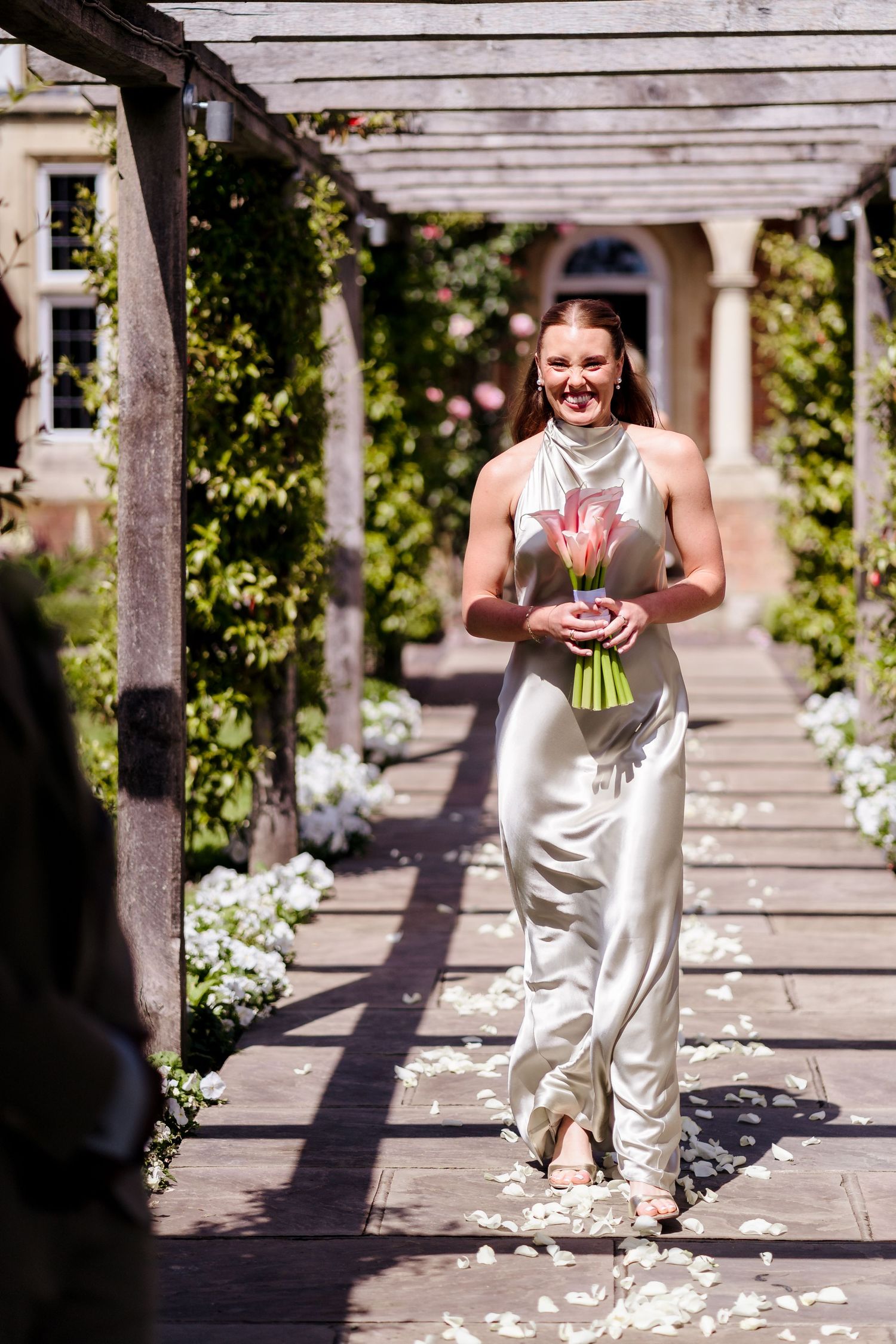 Smiling bride walks down outdoor pergola aisle in silver halter gown holding pink tulip bouquet.
