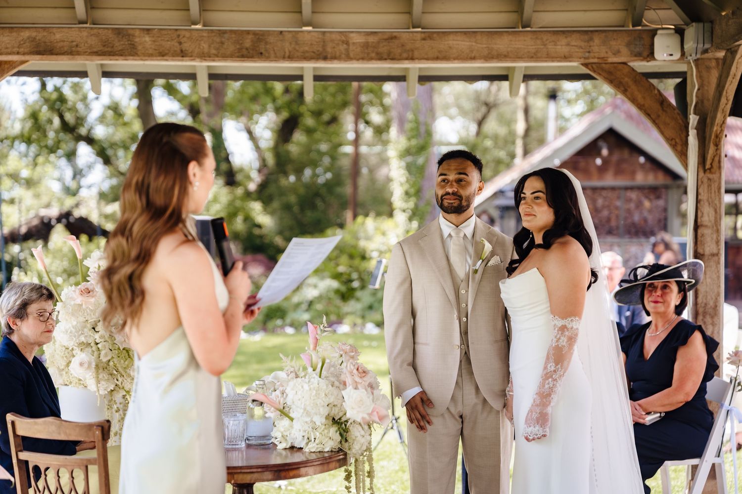 Outdoor wedding ceremony under a pavilion with bride and groom exchanging vows as officiant reads from a book.