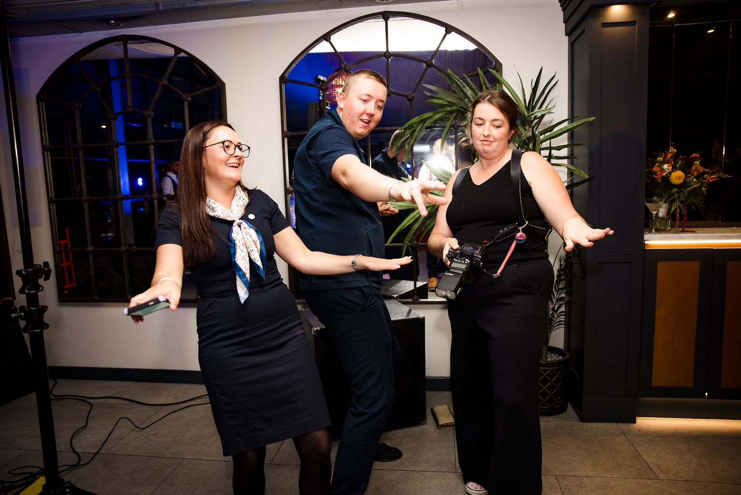Three people dancing and celebrating at an indoor evening event, holding champagne glasses in a stylish venue.