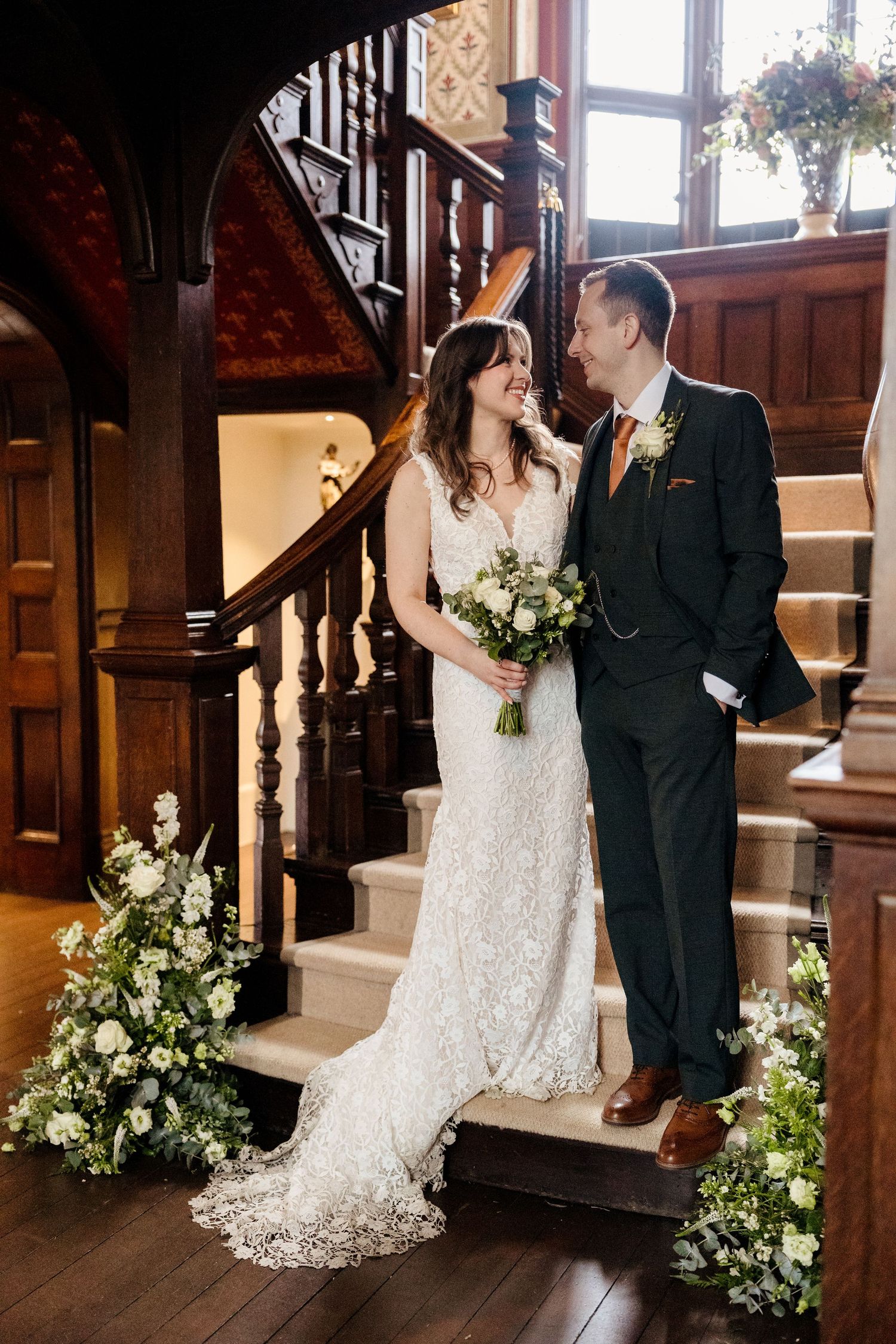 Bride in lace gown and groom in dark suit pose on grand wooden staircase with white floral arrangements.