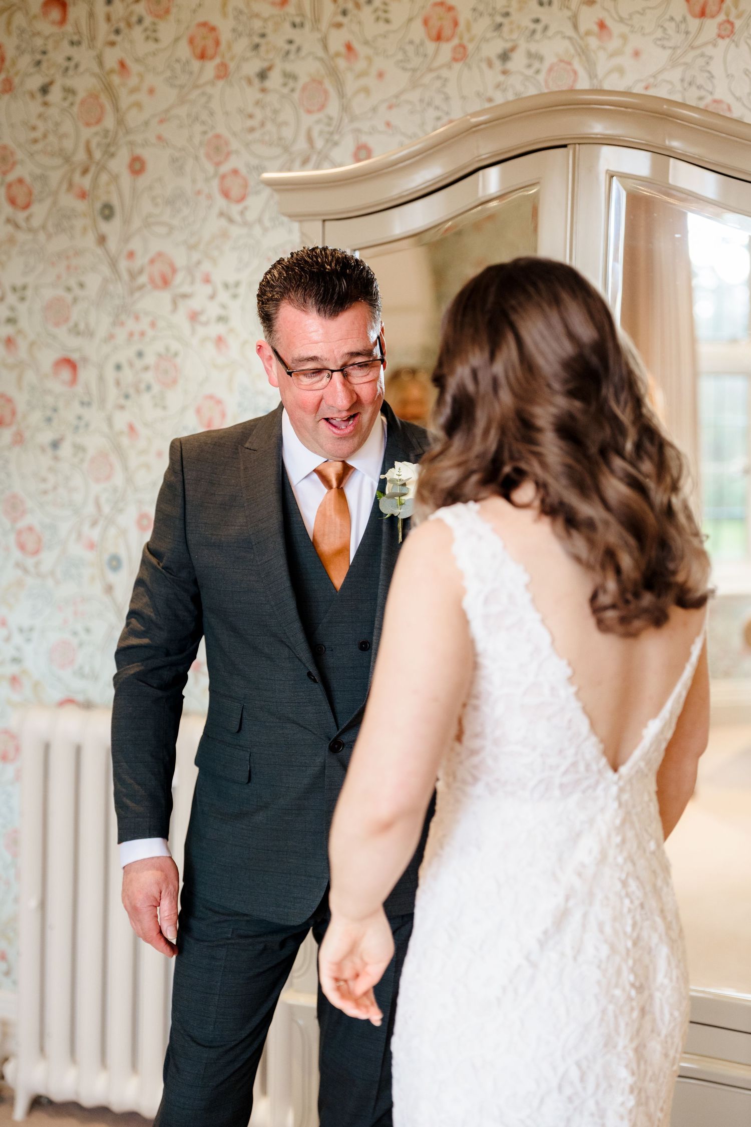Emotional groom smiling at bride during first look in floral wallpapered room wearing dark suit and orange tie.