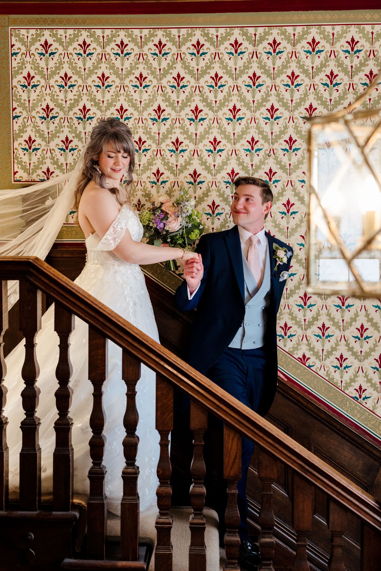 Bride and groom sharing a joyful moment on a wooden staircase with vintage floral wallpaper backdrop.
