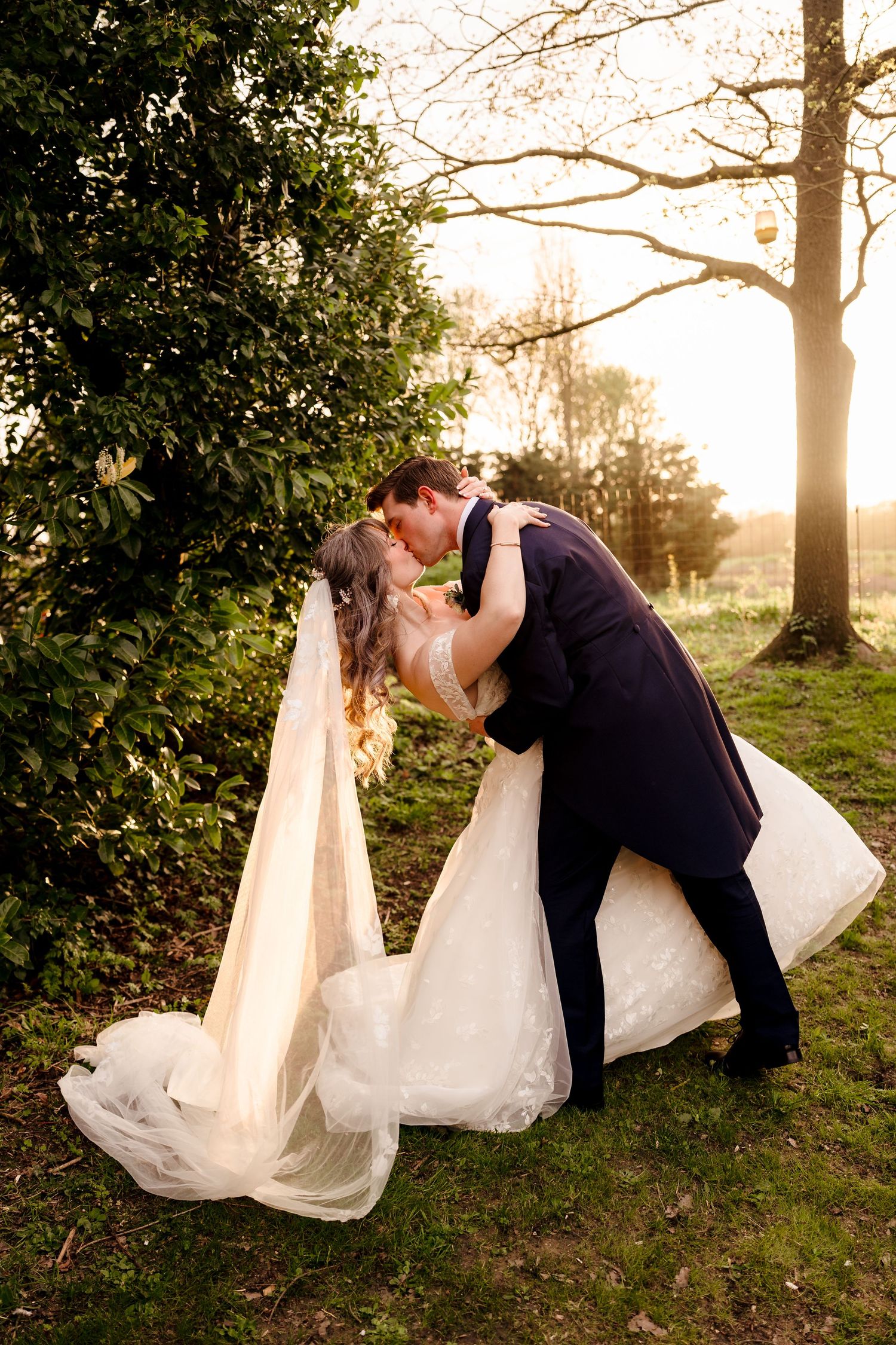Bride and groom sharing romantic dip kiss in golden sunset light beside lush green trees.