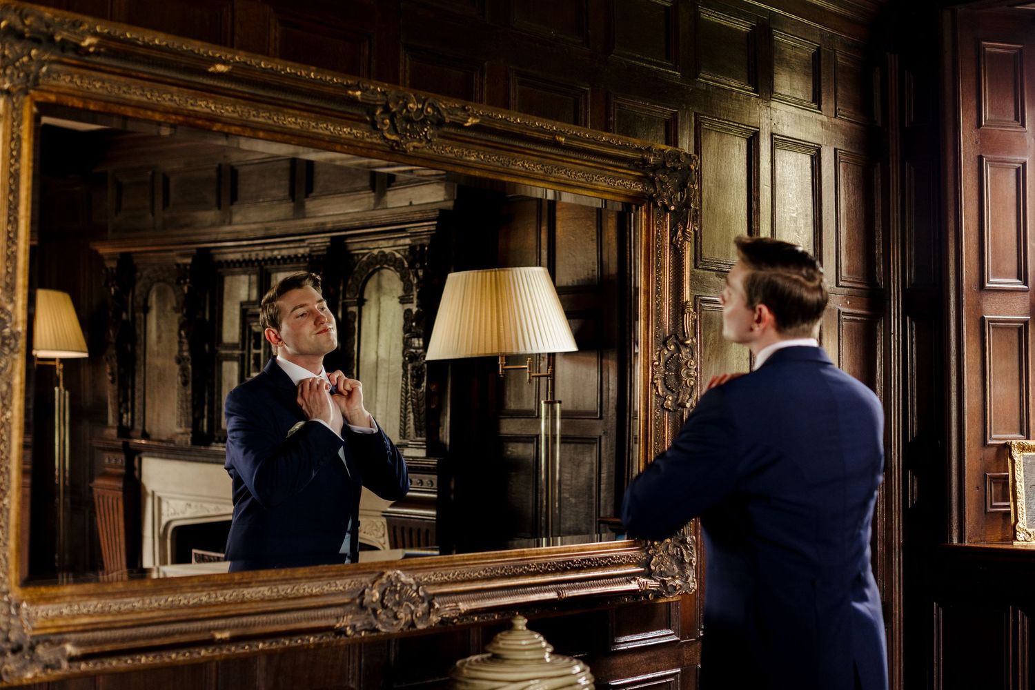 A man in a navy suit adjusts his tie while standing before an ornate gold-framed mirror in an elegant wood-paneled room.