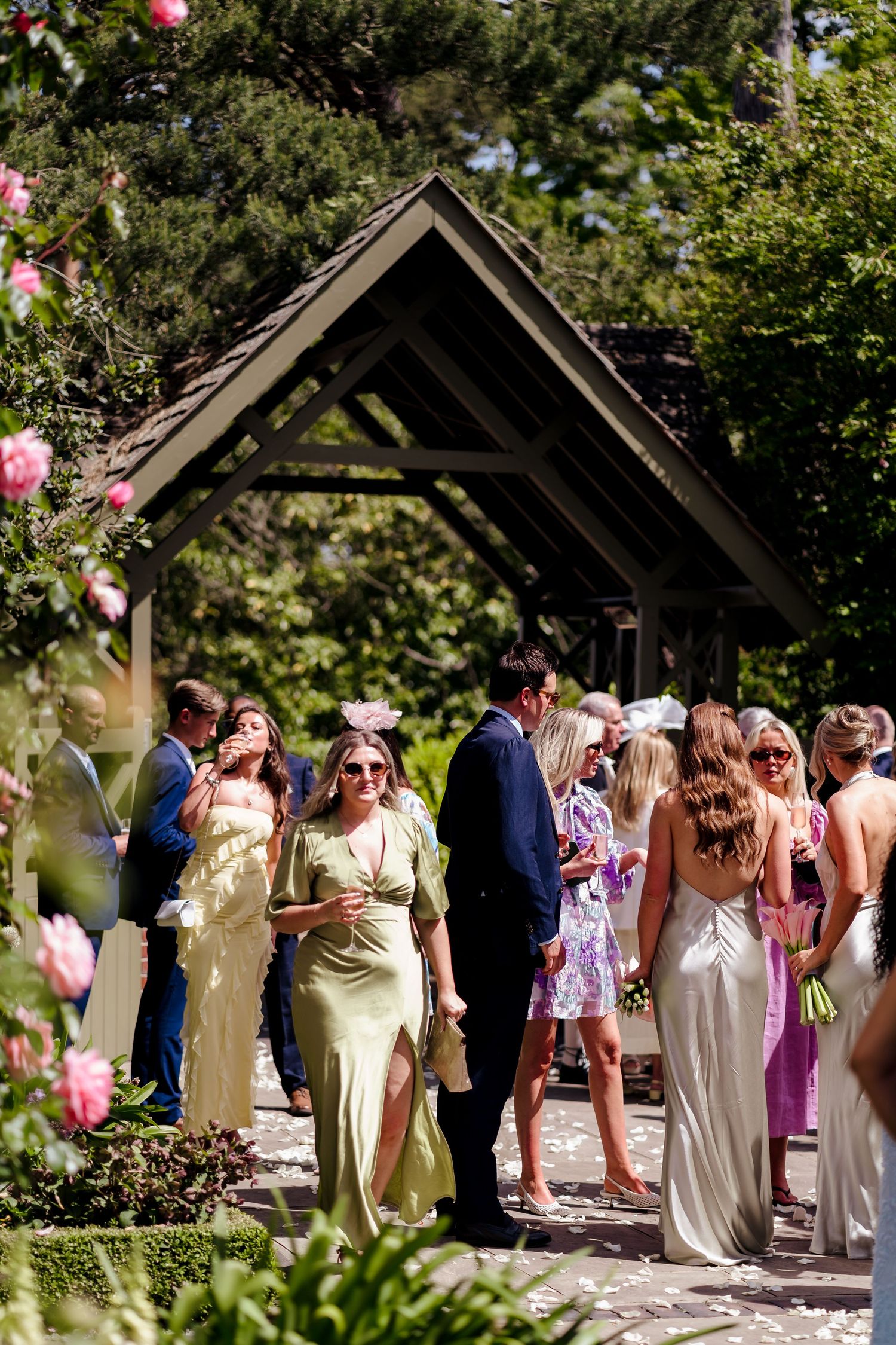 Wedding guests mingling outdoors under a wooden pergola surrounded by lush greenery and roses.