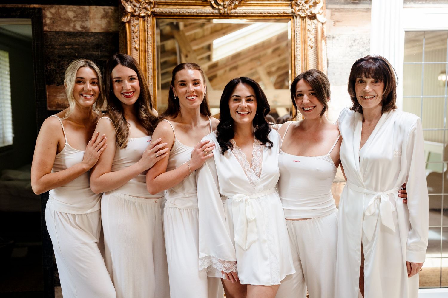Six bridesmaids in white satin robes and slip dresses smiling together before a gold mirror on wedding day.
