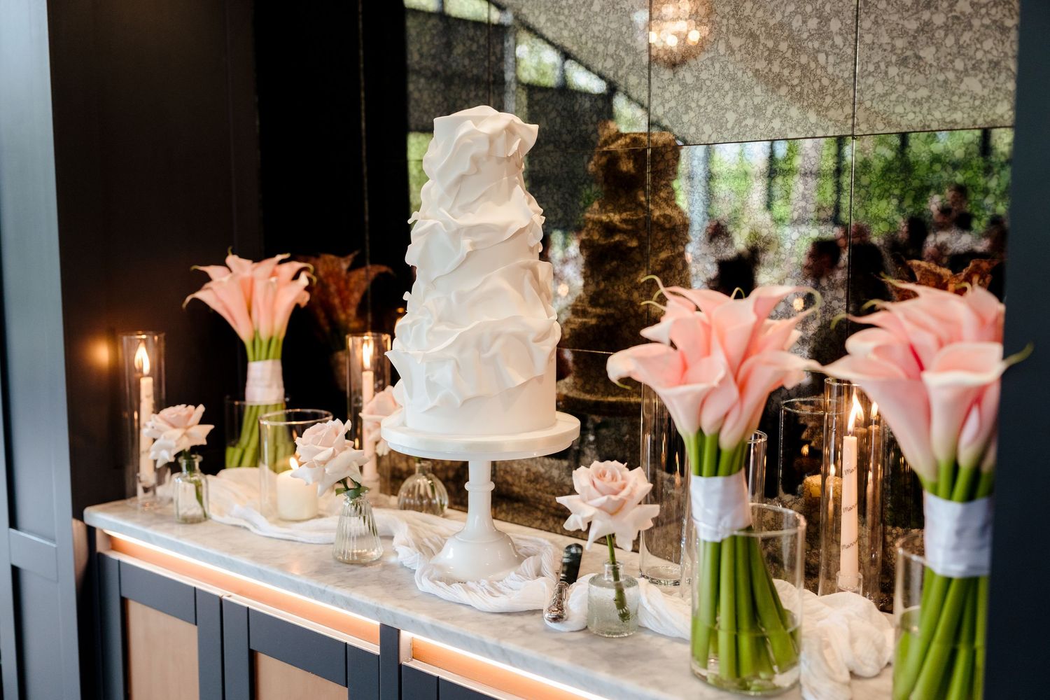 White tiered wedding cake displayed with pink calla lilies and candles on a mirrored table.