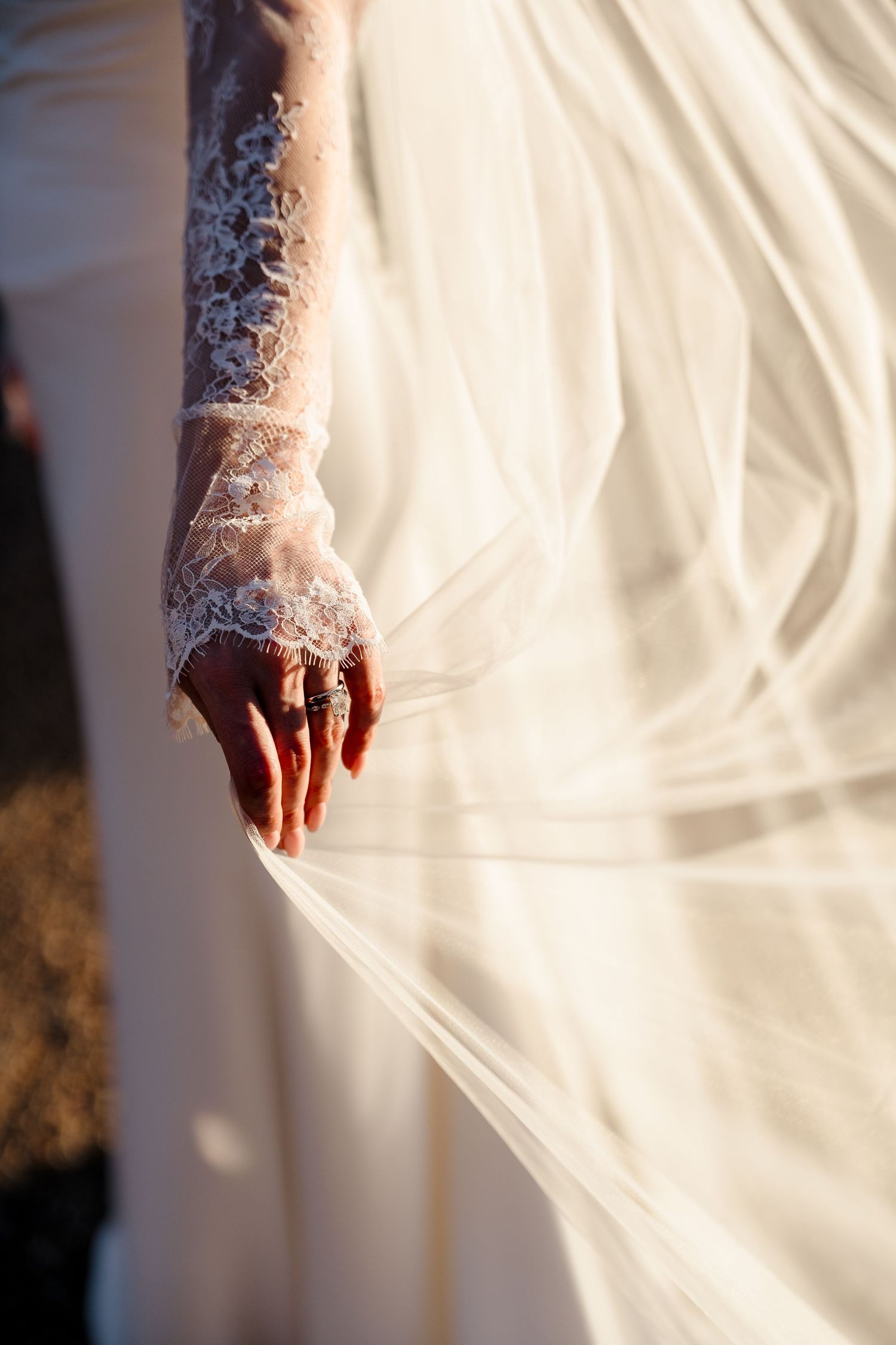 Close-up of a bride's lace-sleeved hand adorned with rings, gently holding her flowing white wedding veil.
