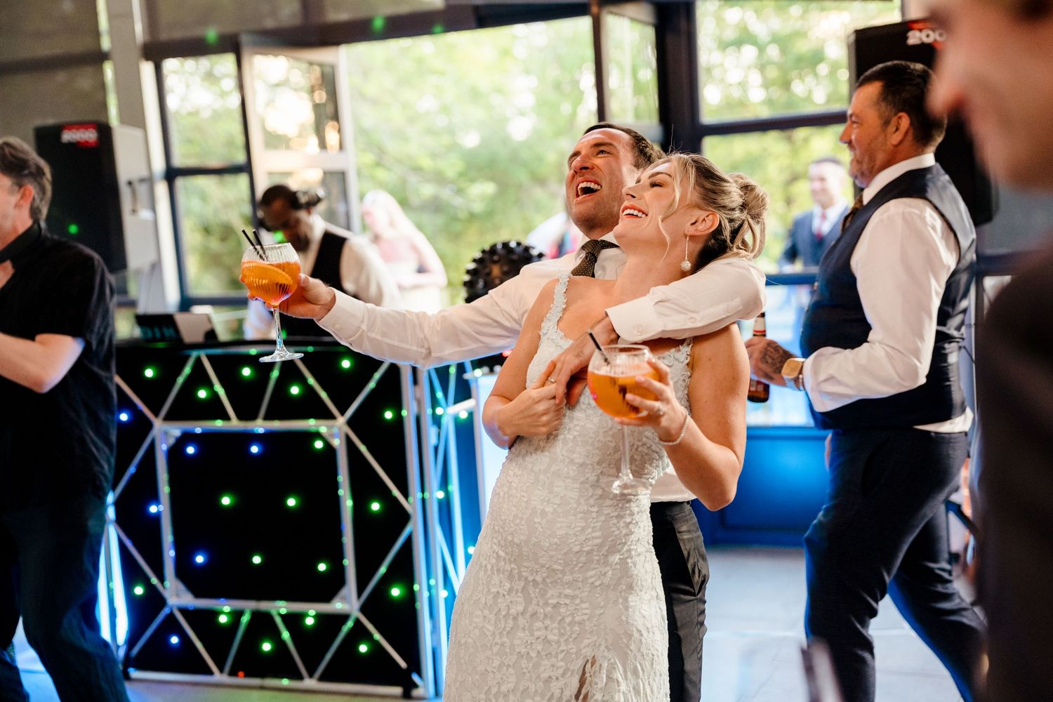 Bride and groom dance joyfully holding cocktails on lit dance floor at lively wedding reception.
