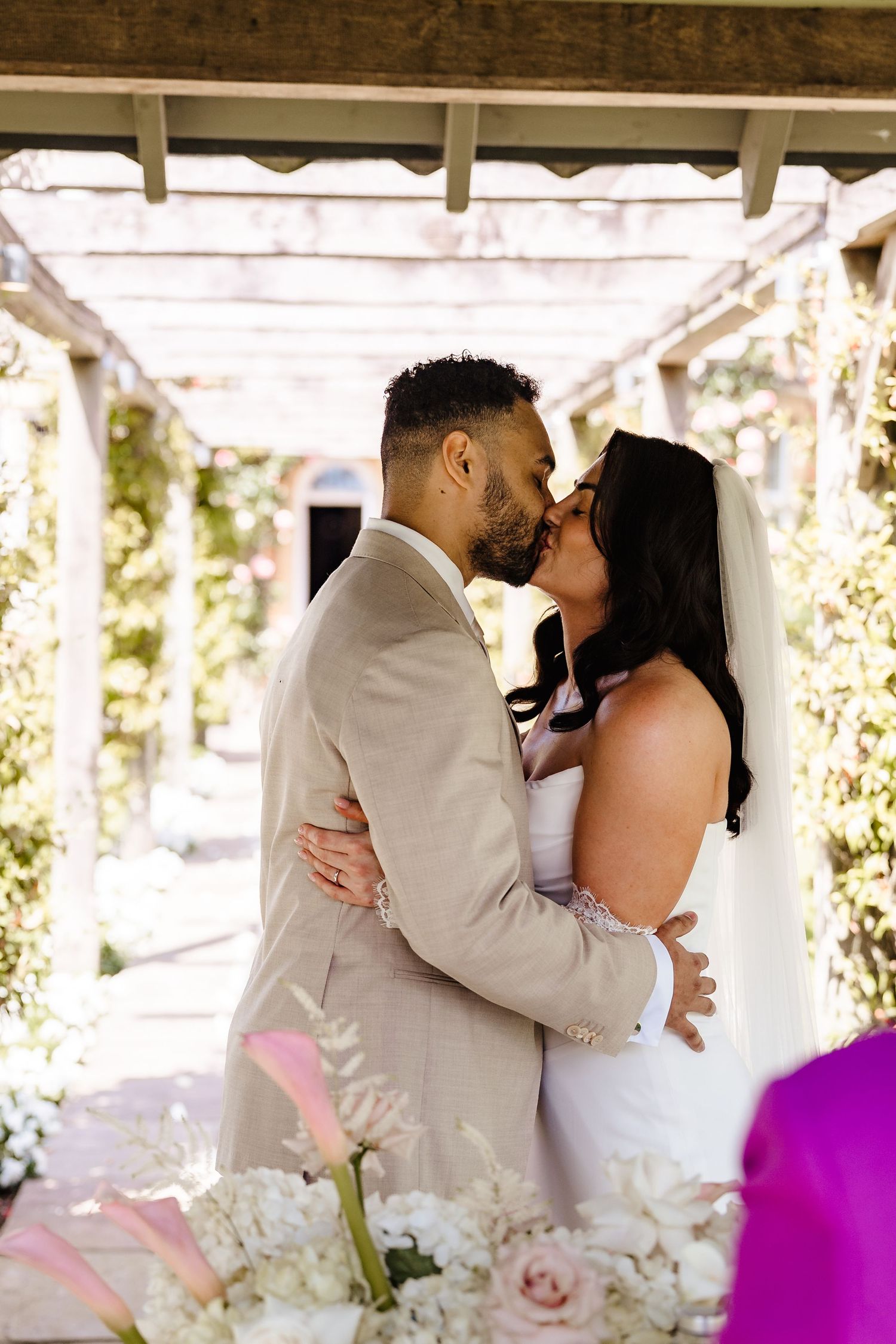 Bride and groom share first kiss under floral pergola during outdoor wedding ceremony.