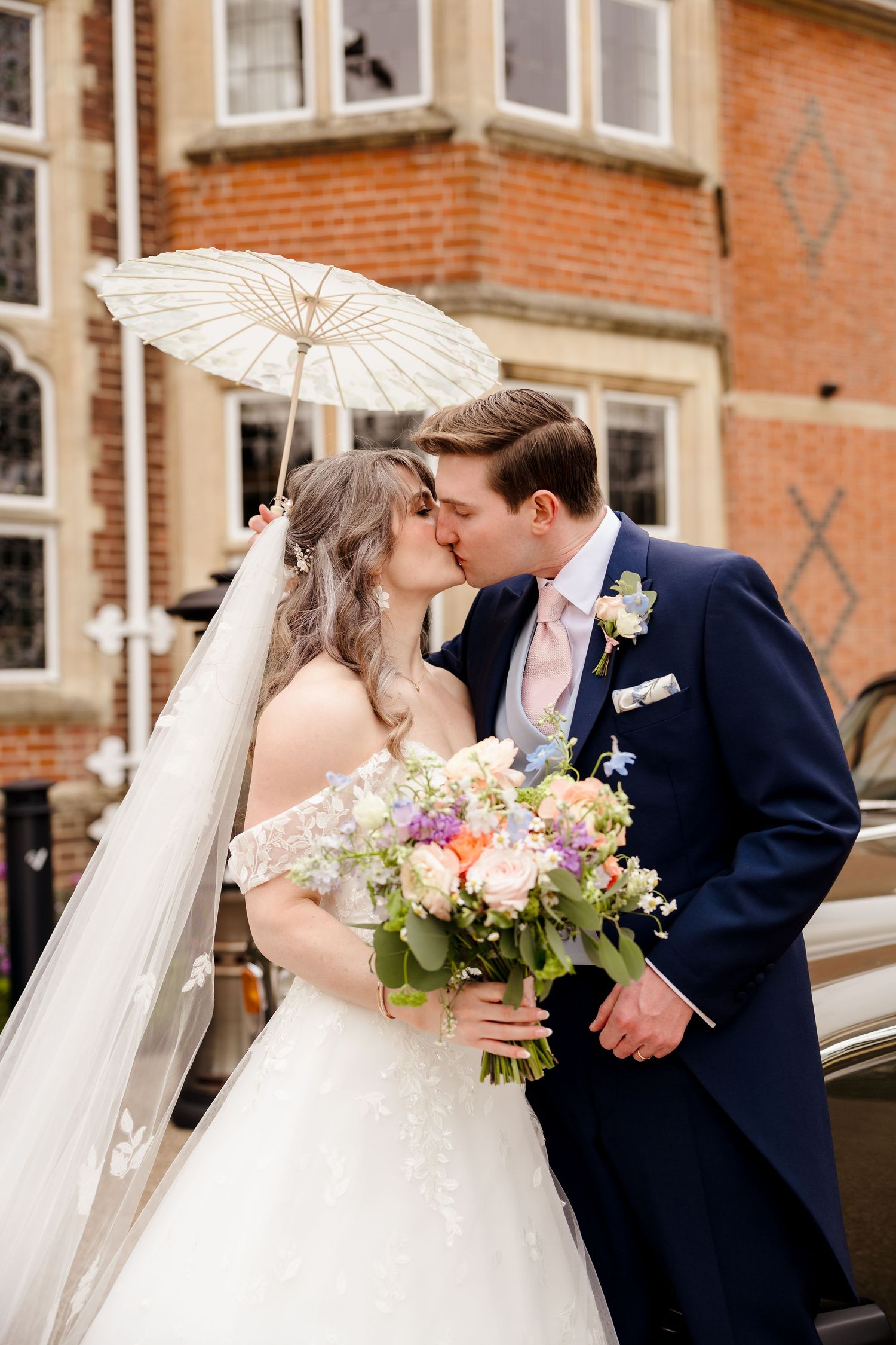 Bride and groom kissing under white parasol outside historic brick building on wedding day.