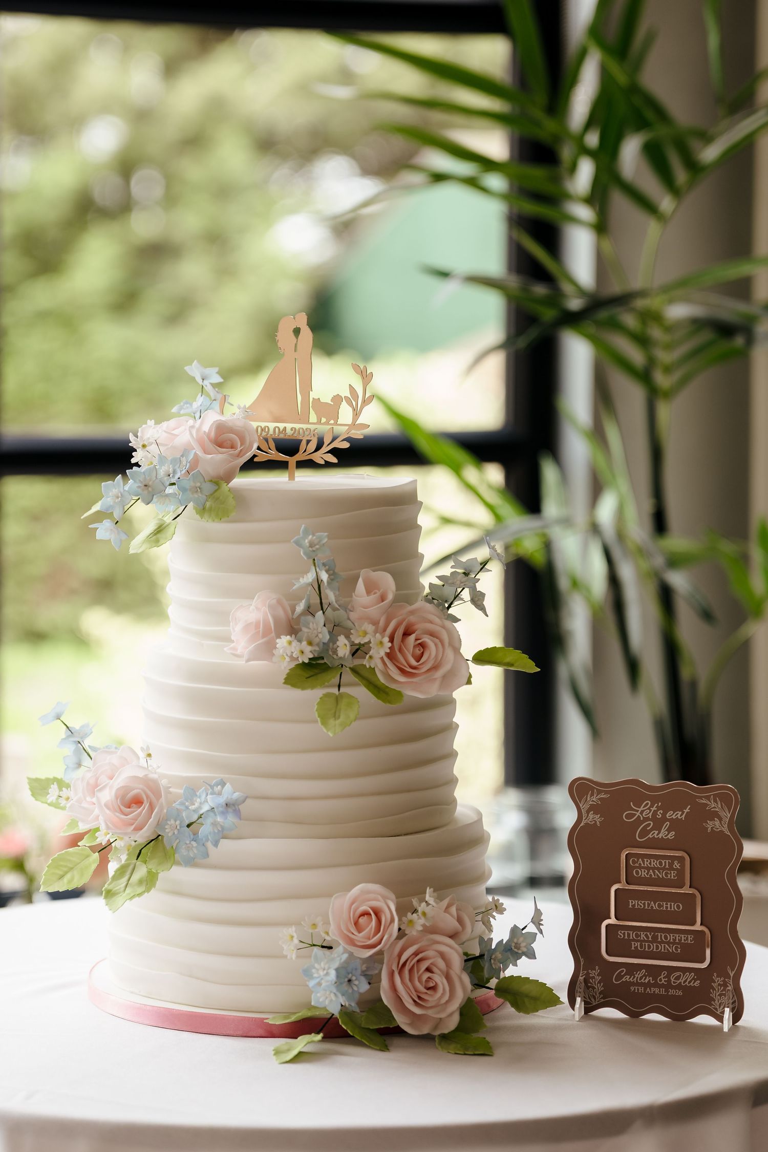 Elegant three-tier white wedding cake with pink roses, blue flowers, and a wooden couple topper on a white table.