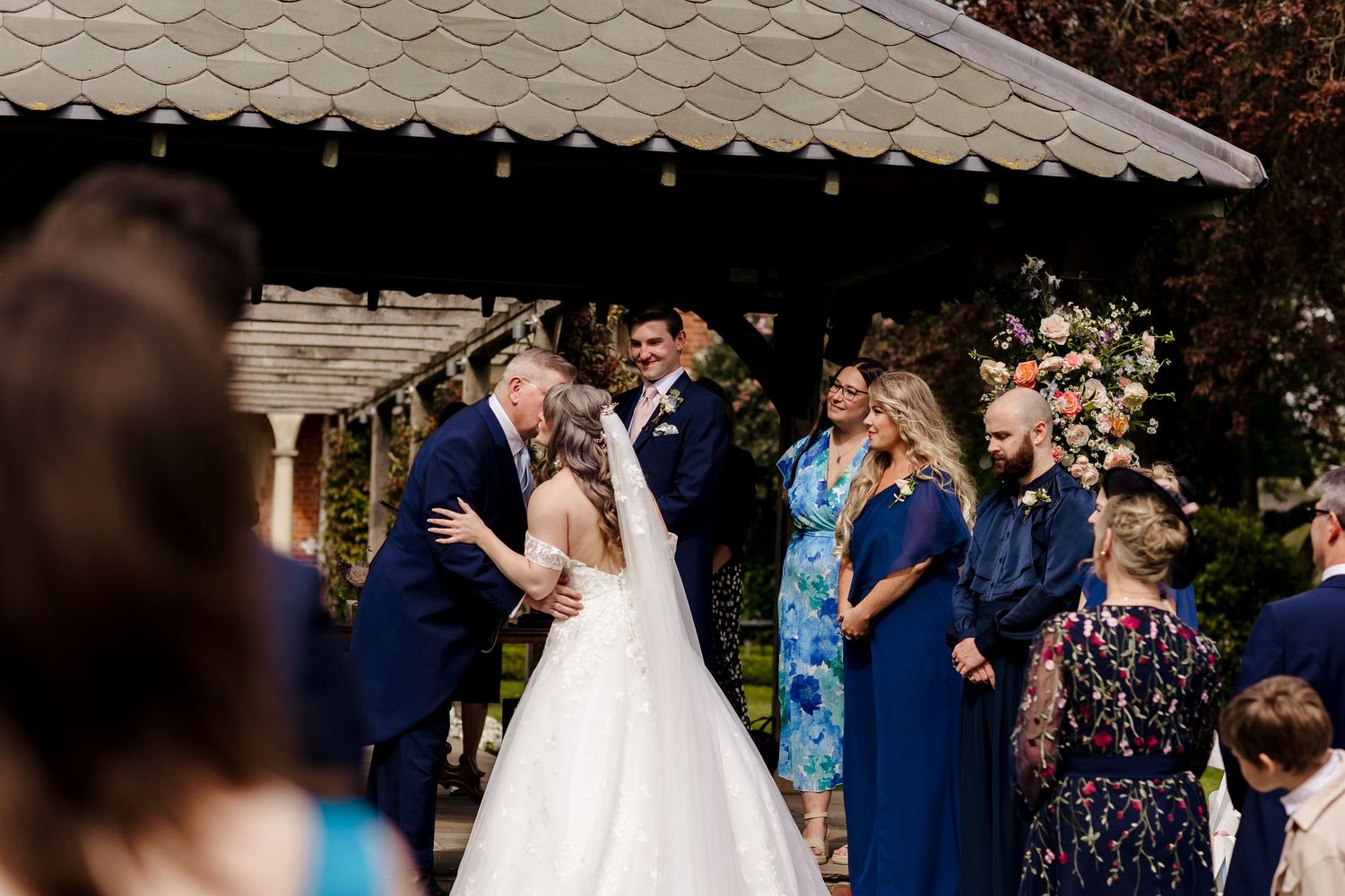 Father of the bride shares a tender kiss with daughter in white gown during outdoor wedding ceremony with guests.