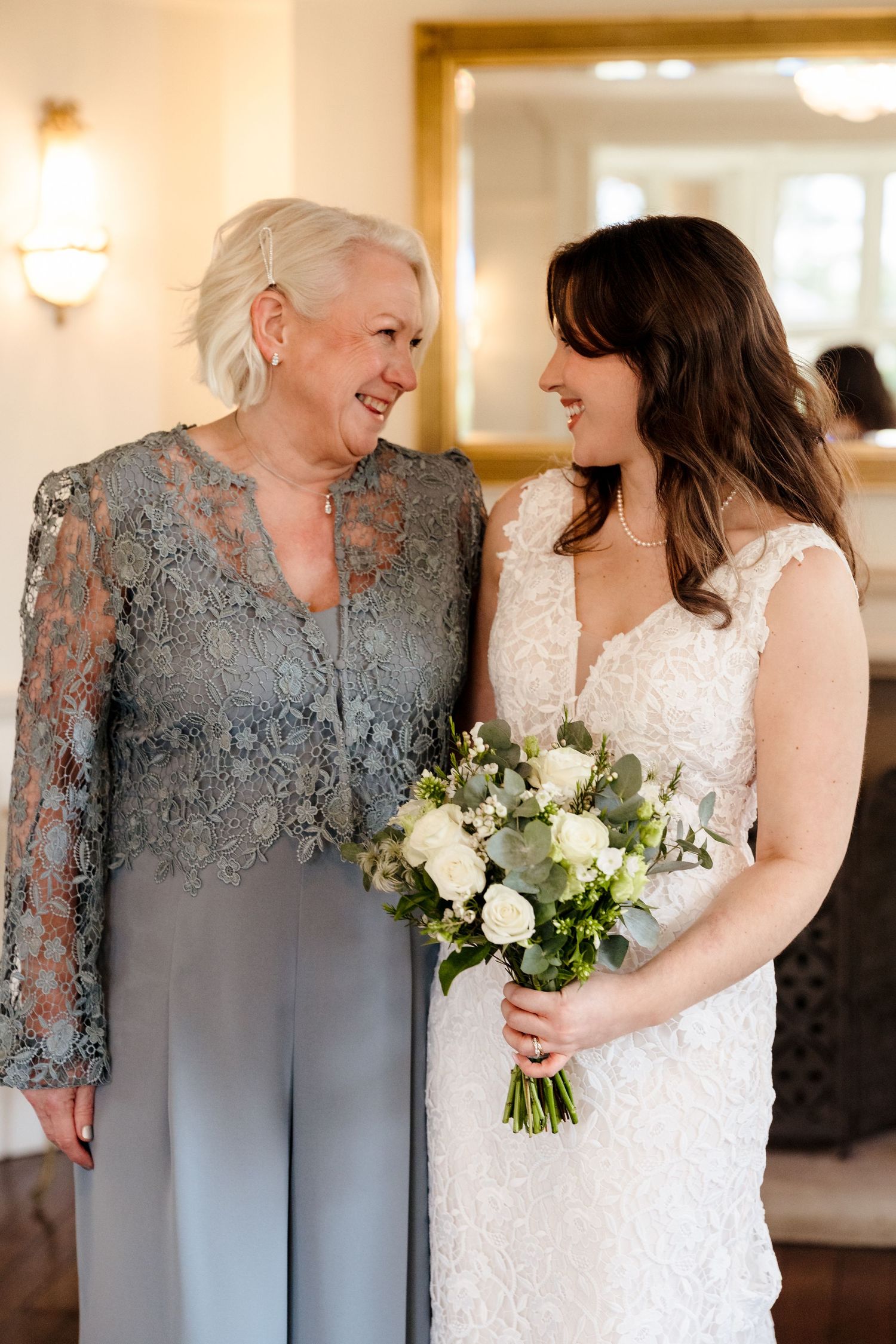 Bride in white lace gown holds green and white bouquet smiling with older woman in blue lace outfit at wedding.