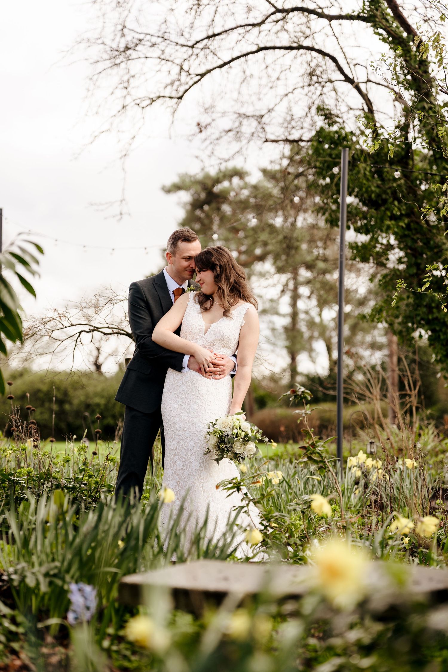 A bride and groom embrace romantically in a lush garden, surrounded by yellow flowers and greenery on their wedding day.