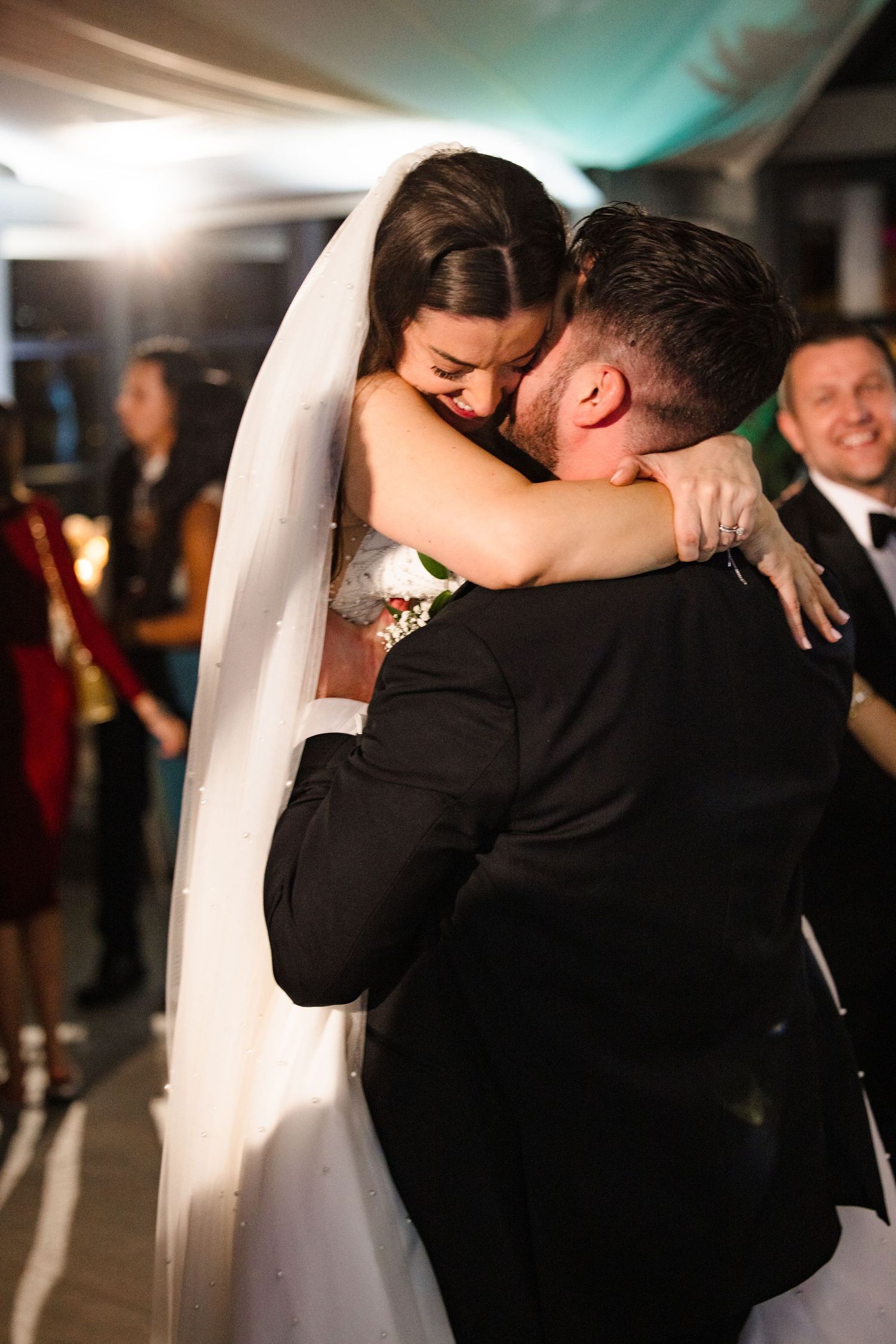 Bride and groom sharing emotional first dance hug at wedding reception, guests celebrating in background.