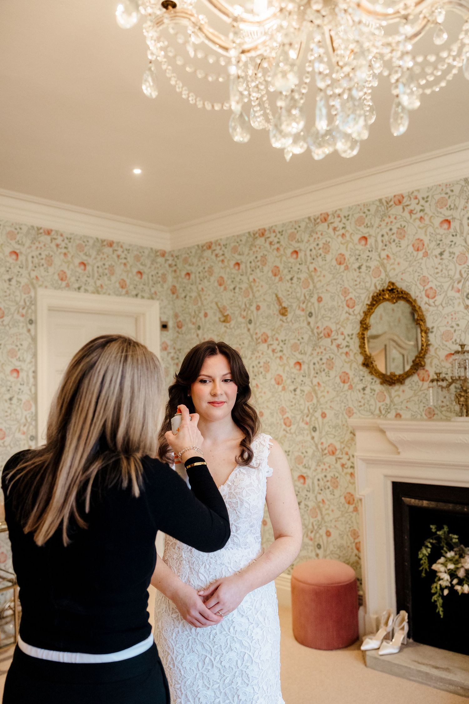 Bride in lace wedding dress gets final touches from makeup artist in elegant floral wallpapered room.