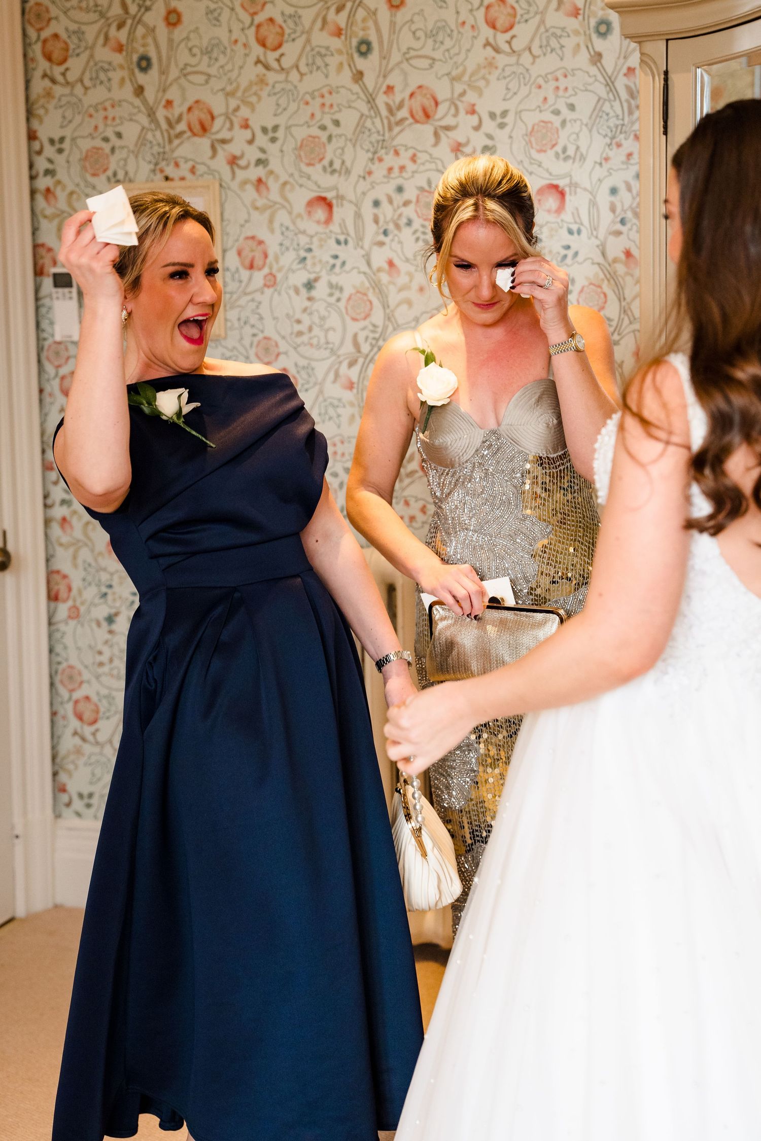 Bridesmaids in navy and gold dresses react emotionally while helping bride prepare for wedding ceremony.