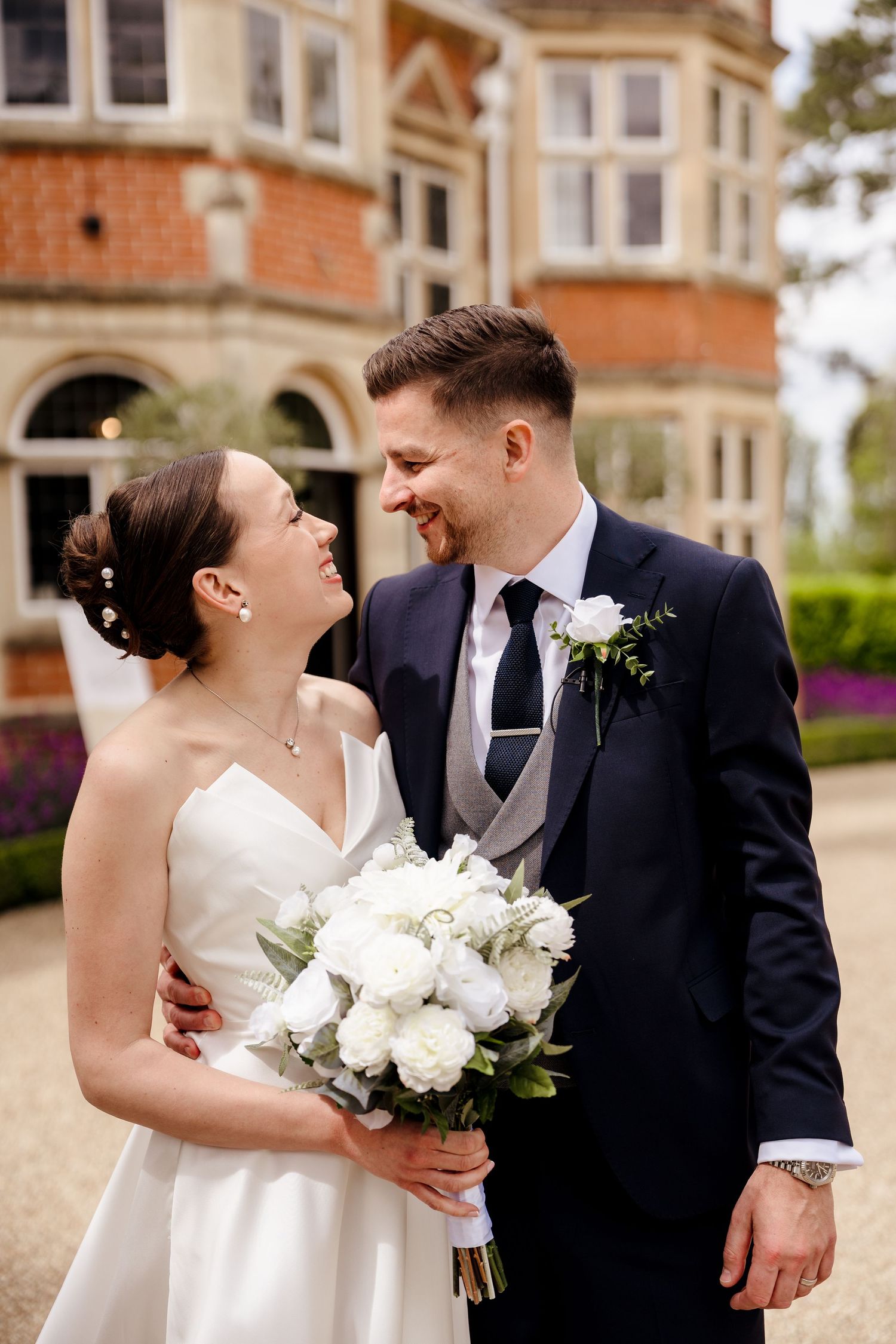 A smiling bride and groom share a romantic moment outside a grand brick manor, bride holding a white peony bouquet.