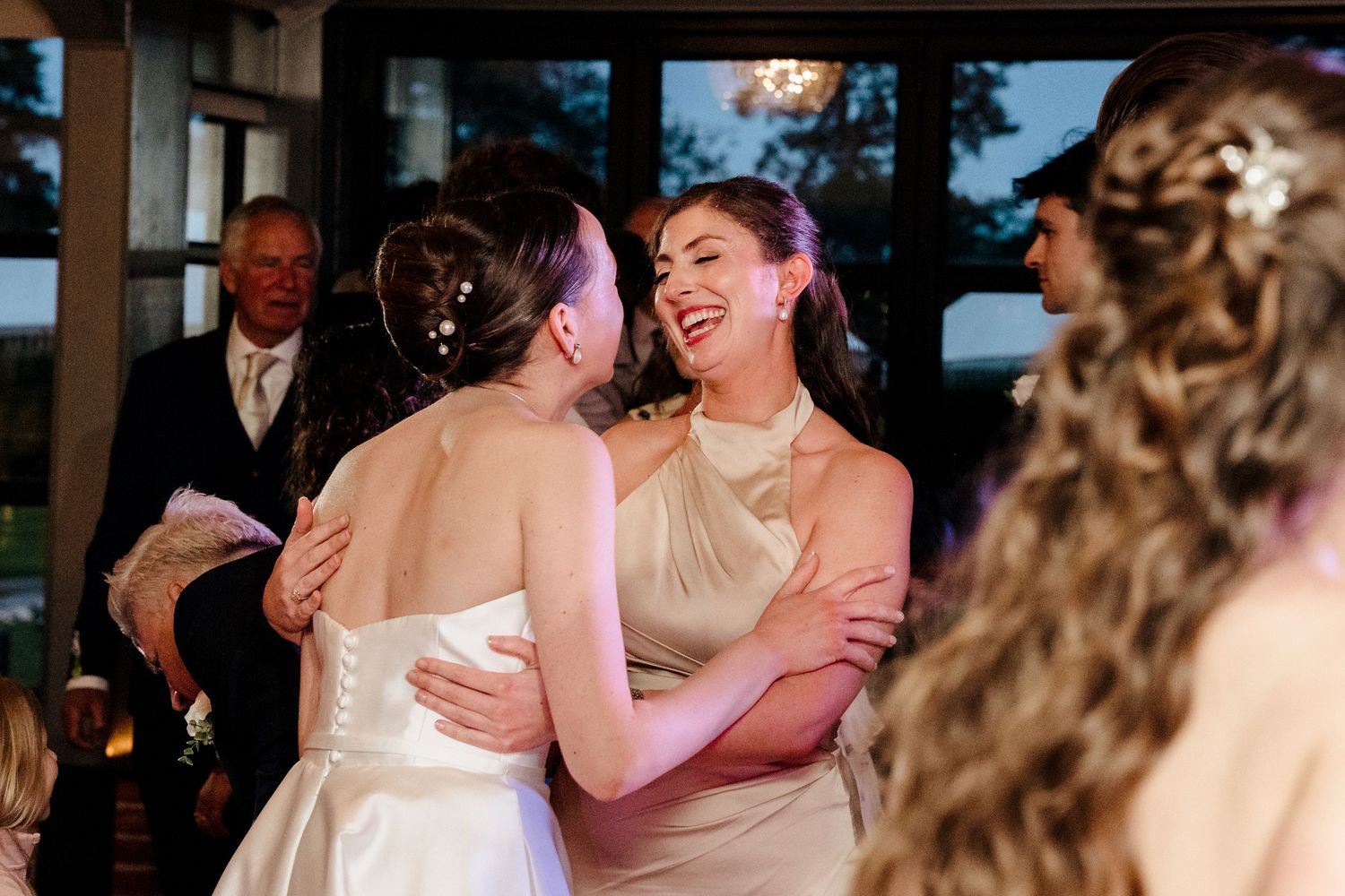 Bride embraces laughing bridesmaid in champagne dress during wedding reception celebration.