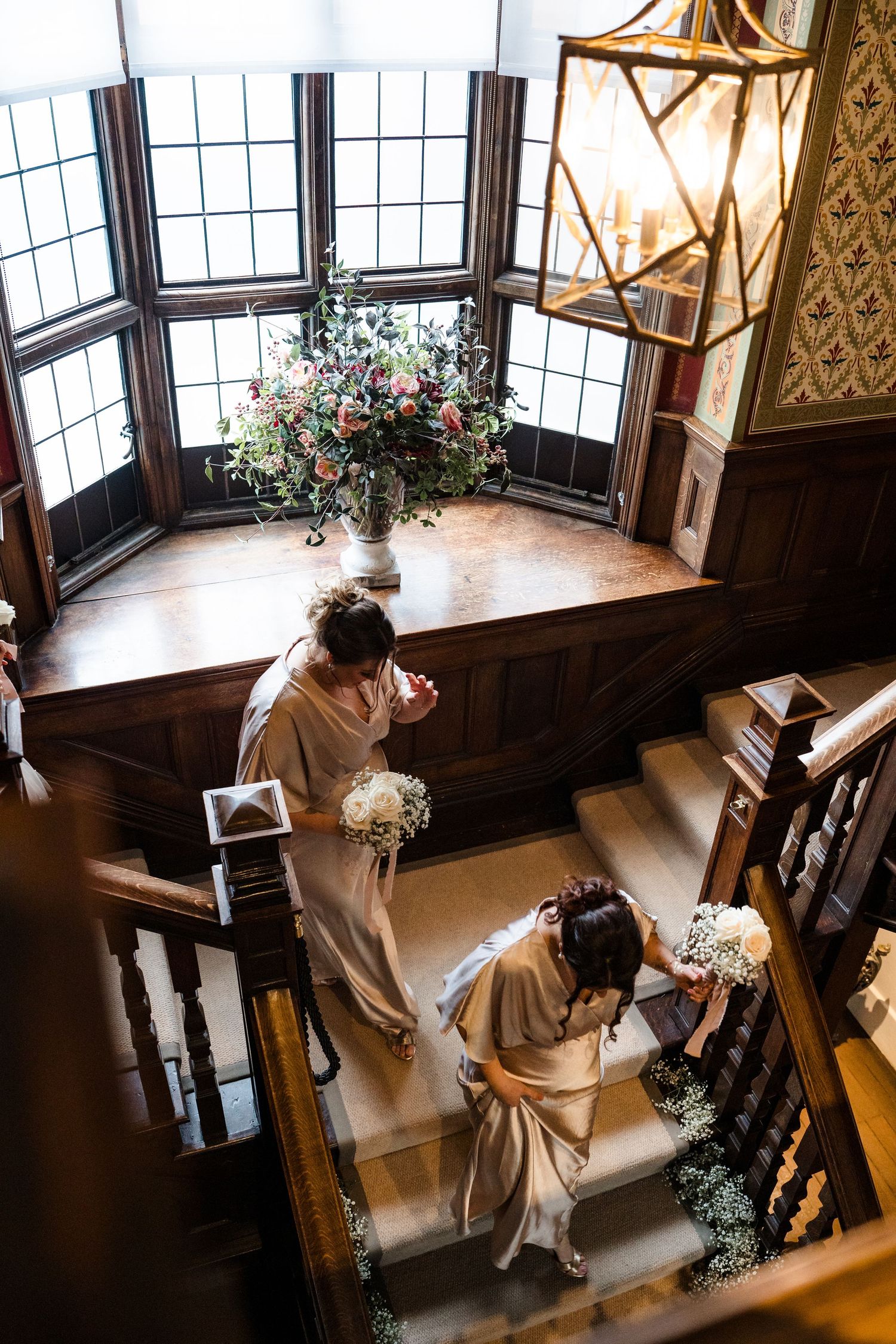 Aerial view of bridesmaids in champagne gowns descending a grand wooden staircase with floral bouquets.