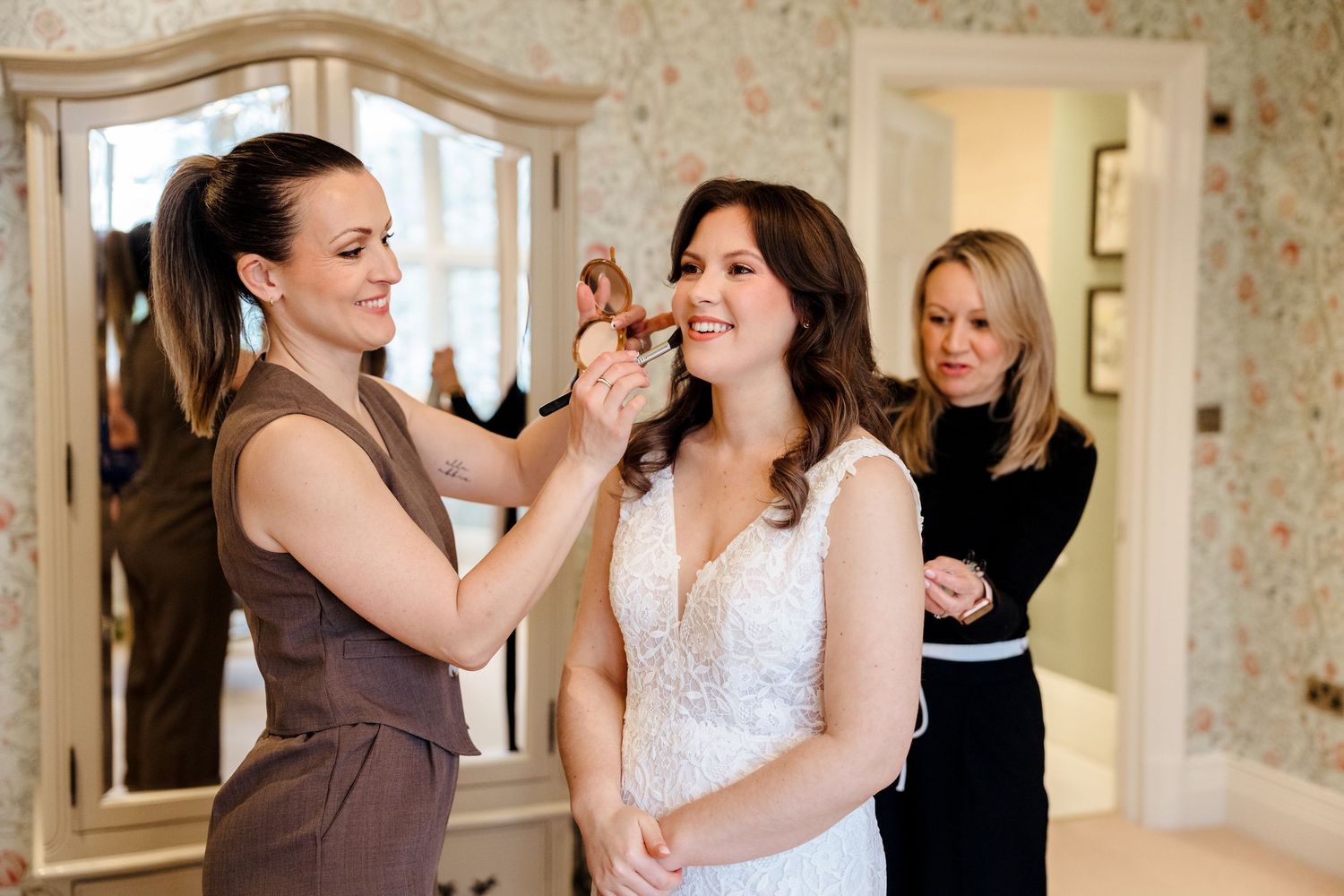 Bride in white lace gown gets makeup applied by stylist while another woman assists in elegant floral room.