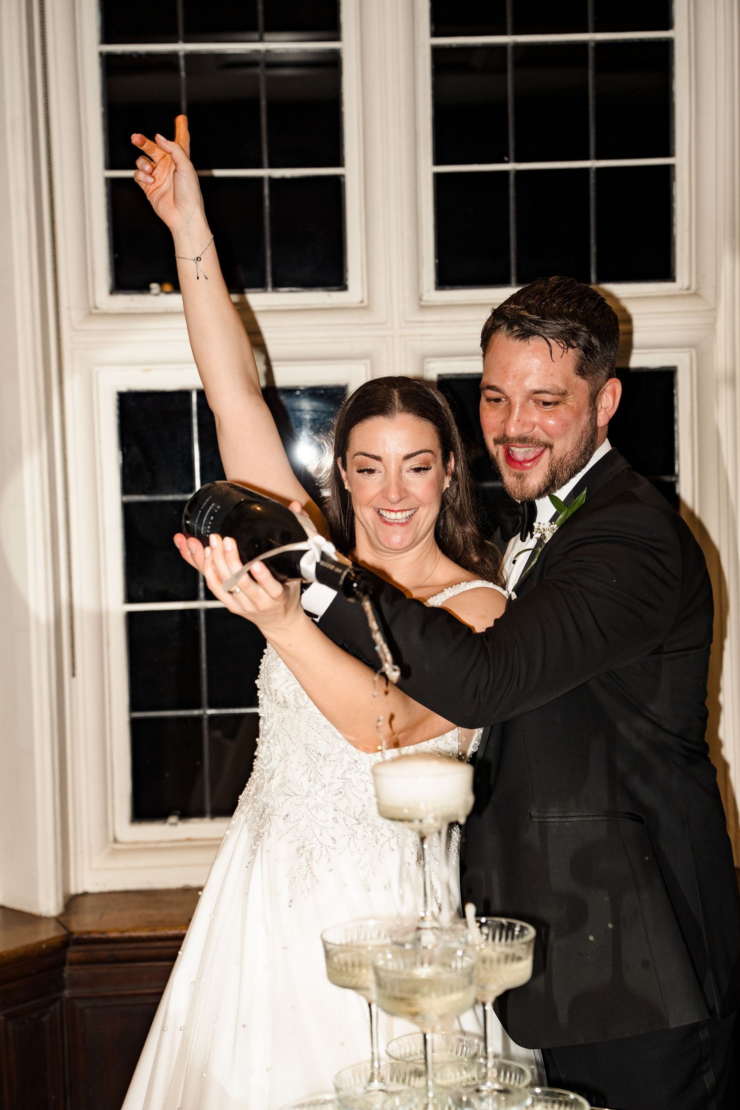 Bride and groom joyfully pour champagne tower together at their elegant evening wedding reception.