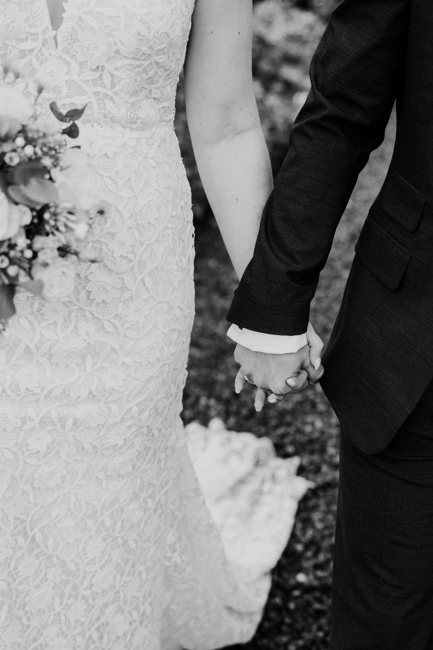 Black and white photo of bride and groom holding hands, bride in lace gown holding bouquet.