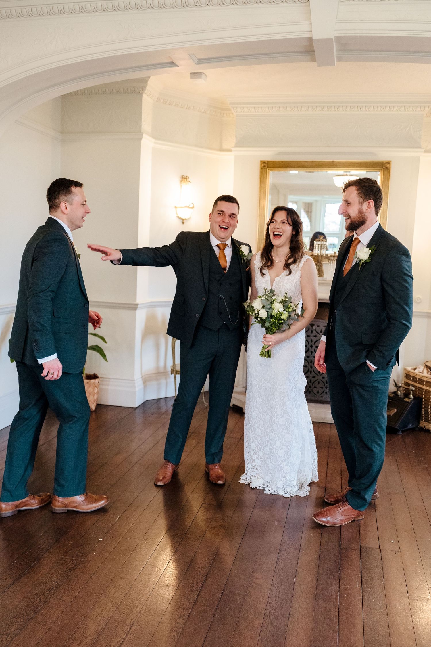 Bride and groom laughing with groomsmen in dark suits and orange ties in an elegant wedding venue.