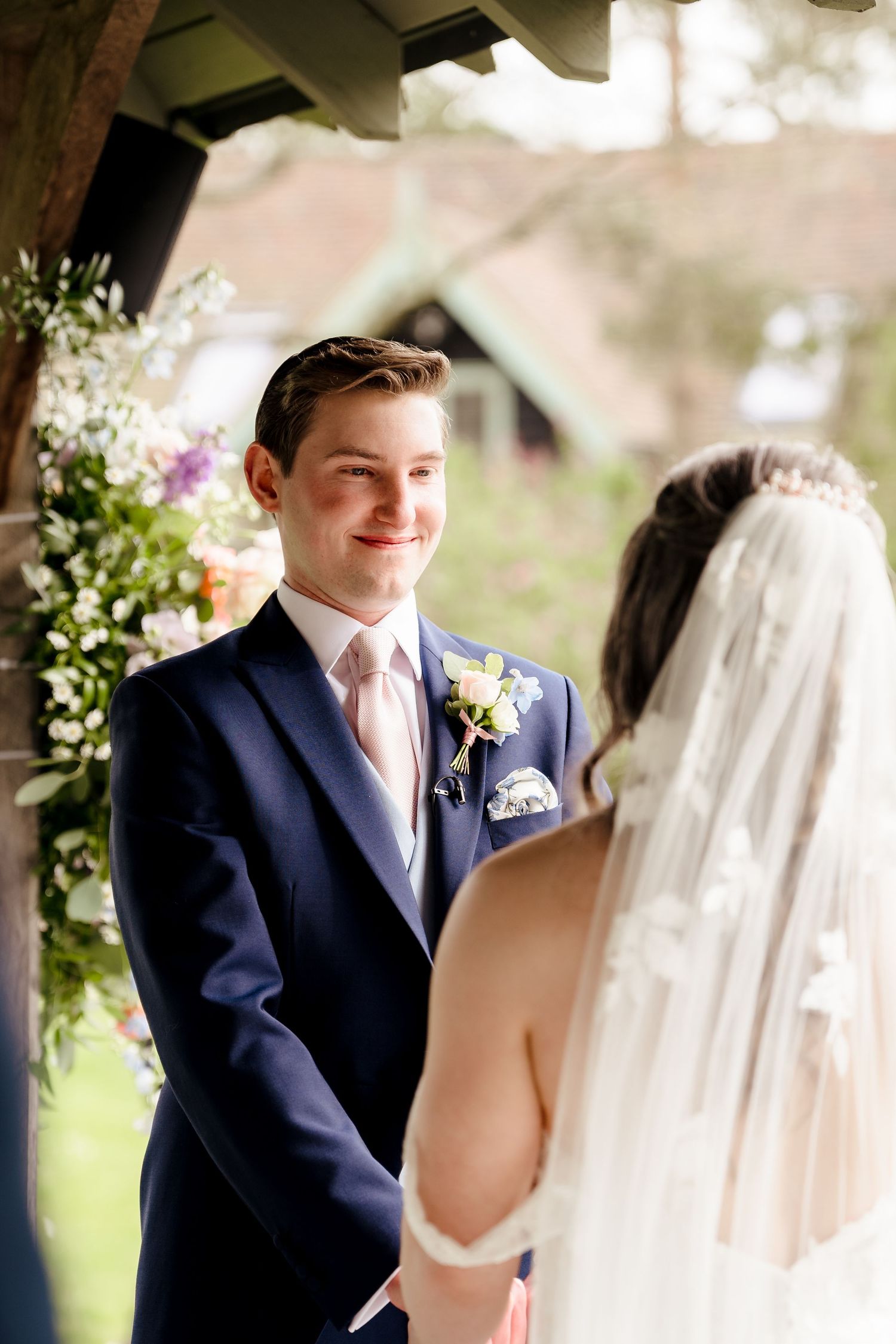 Groom in navy suit smiling tenderly at bride in veil during outdoor wedding ceremony.